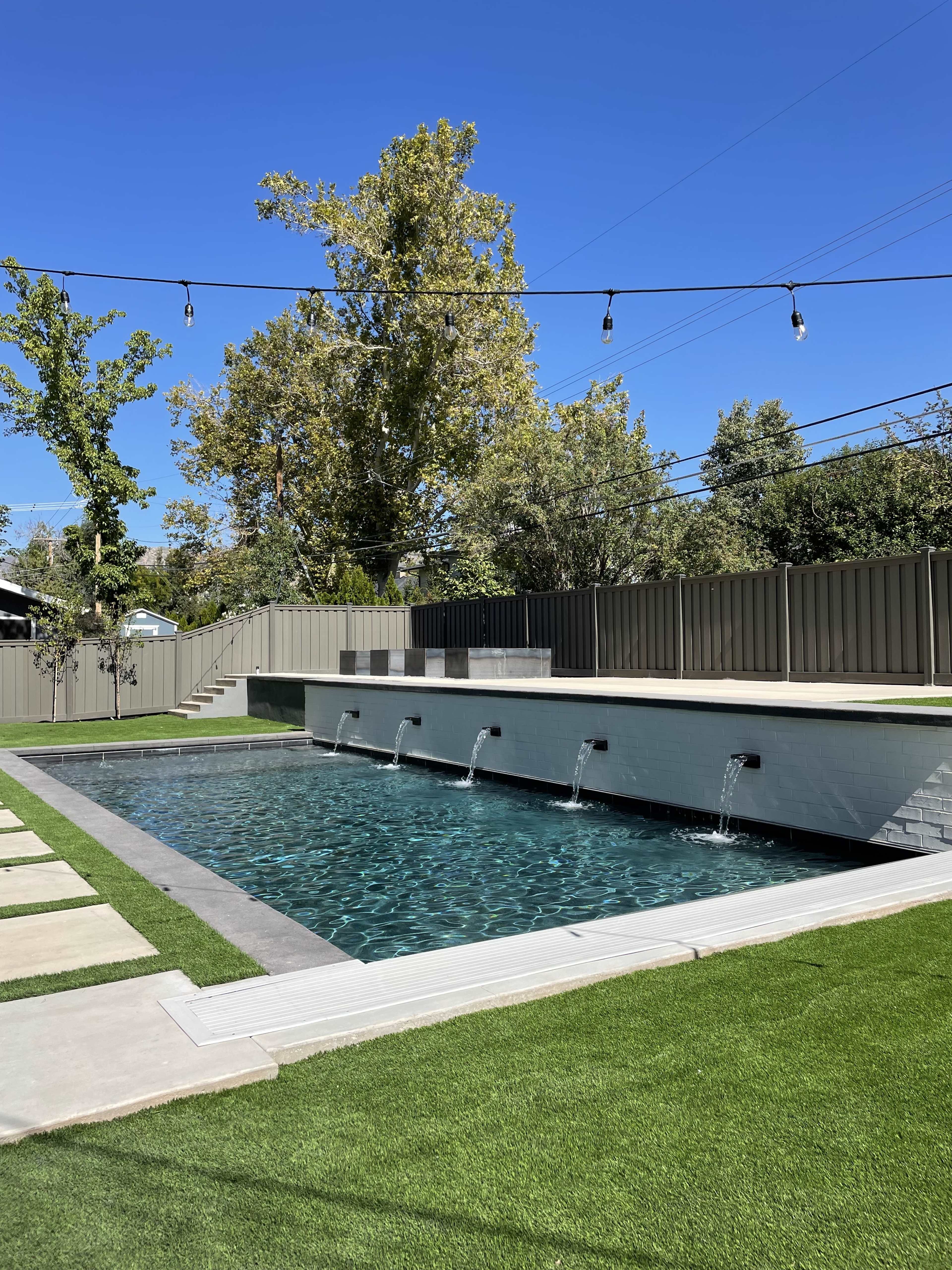 A modern pool surrounded by landscaped grass, with three water spouts along the pool's edge and a wooden fence in the background under a clear blue sky.