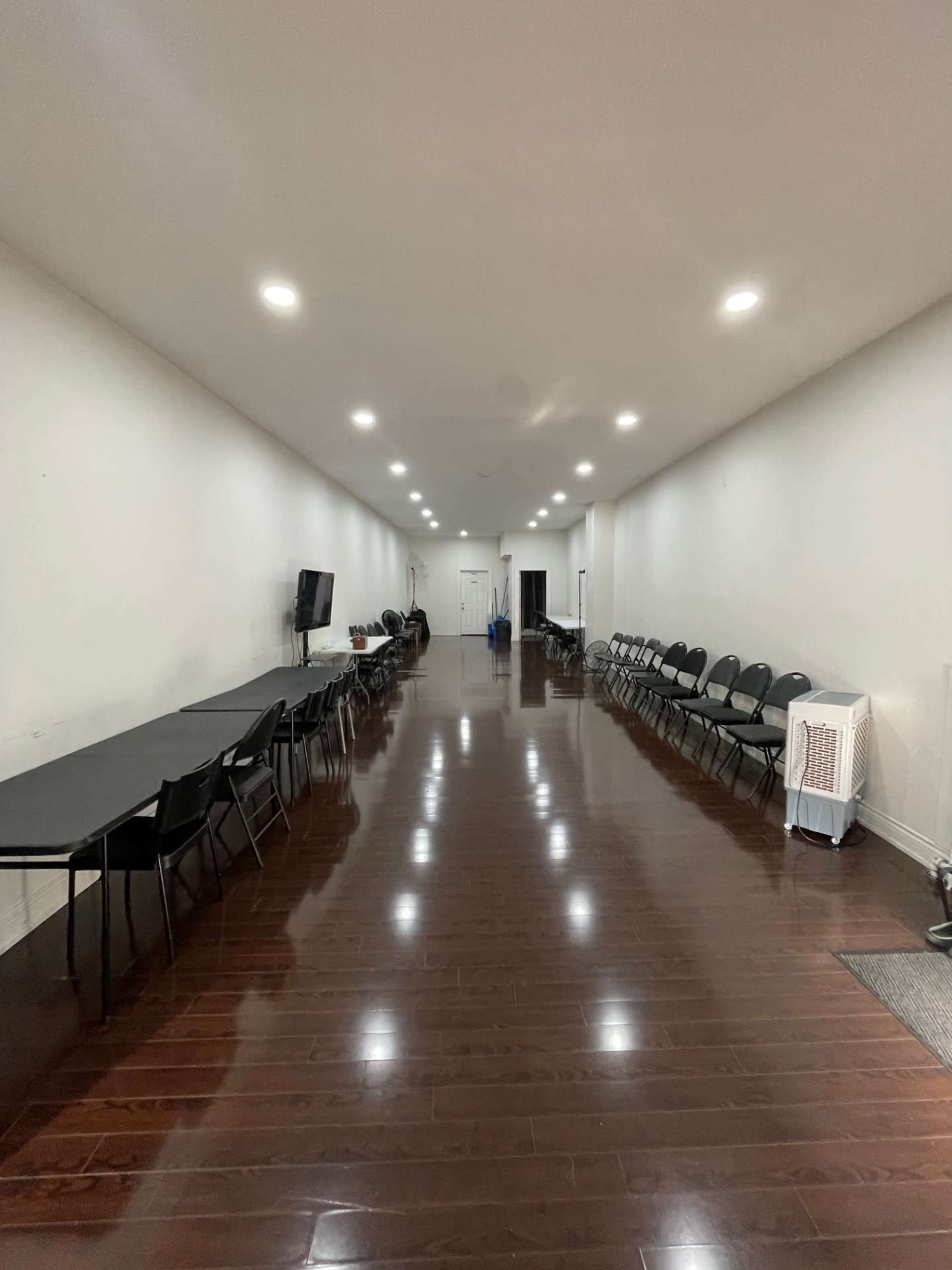 The image shows a long, narrow room with polished wooden floors and rows of black chairs along the sides, with a few tables set up in the center and a television on one wall.