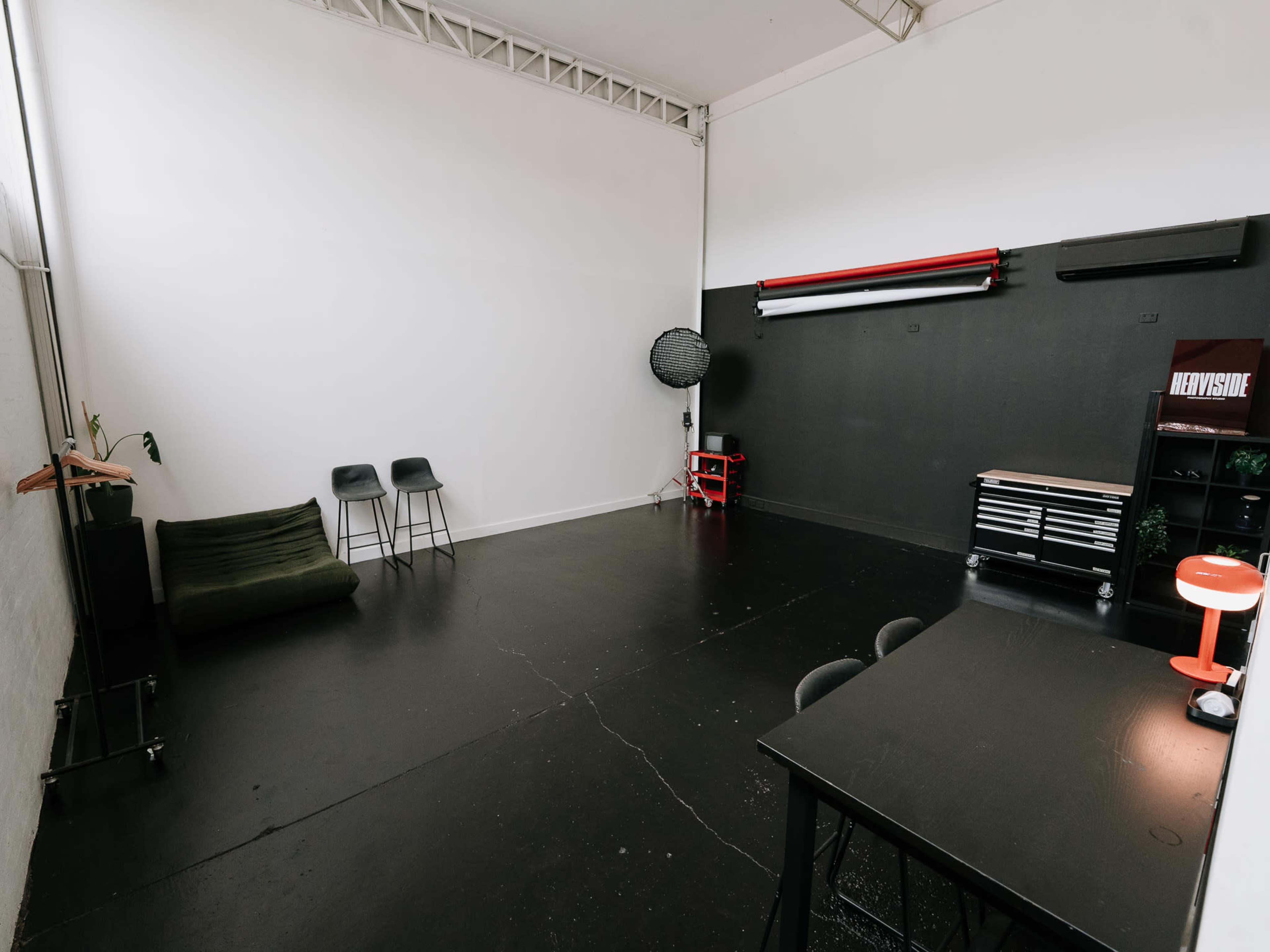The image shows a minimalist studio space with black flooring, a white wall, and a mix of furniture including chairs, a small table, a red lamp, and various equipment against the back wall.