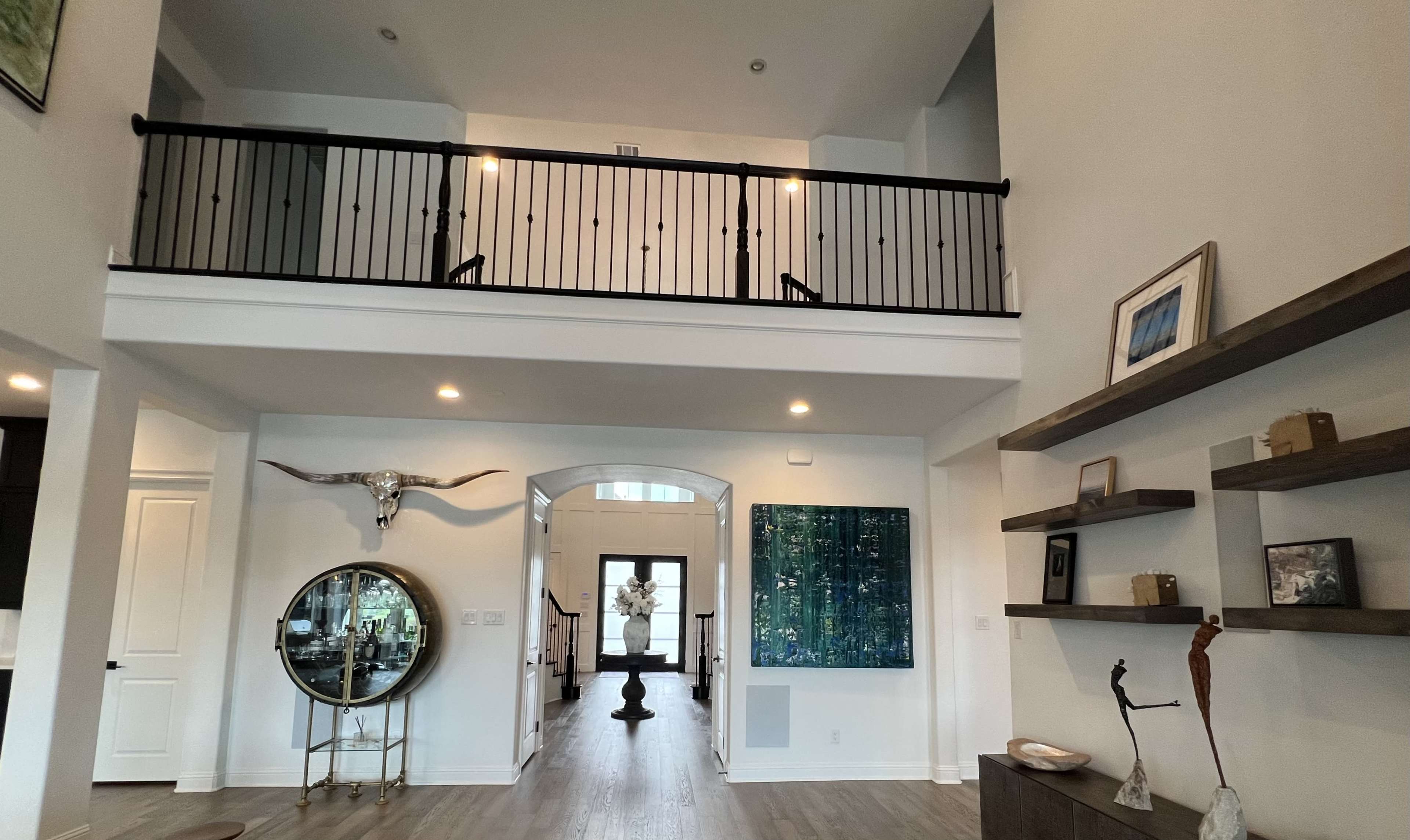 The image shows an interior view of a two-story foyer with a circular mirror, a large green artwork, and wooden shelves, leading up to a balcony with black railings.