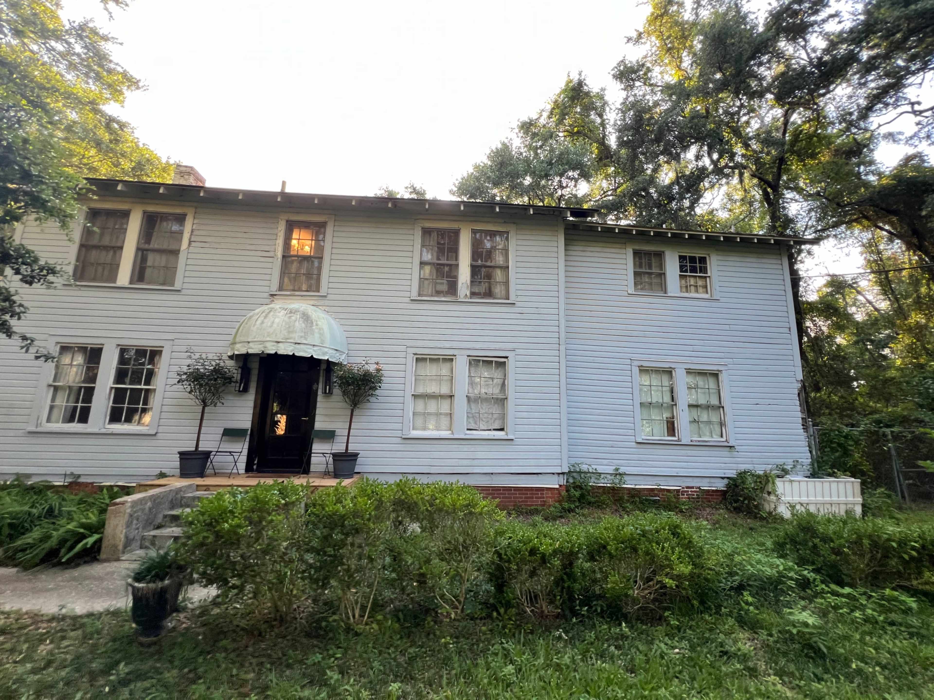 A two-story light blue house with a front entrance featuring an awning and surrounded by greenery.