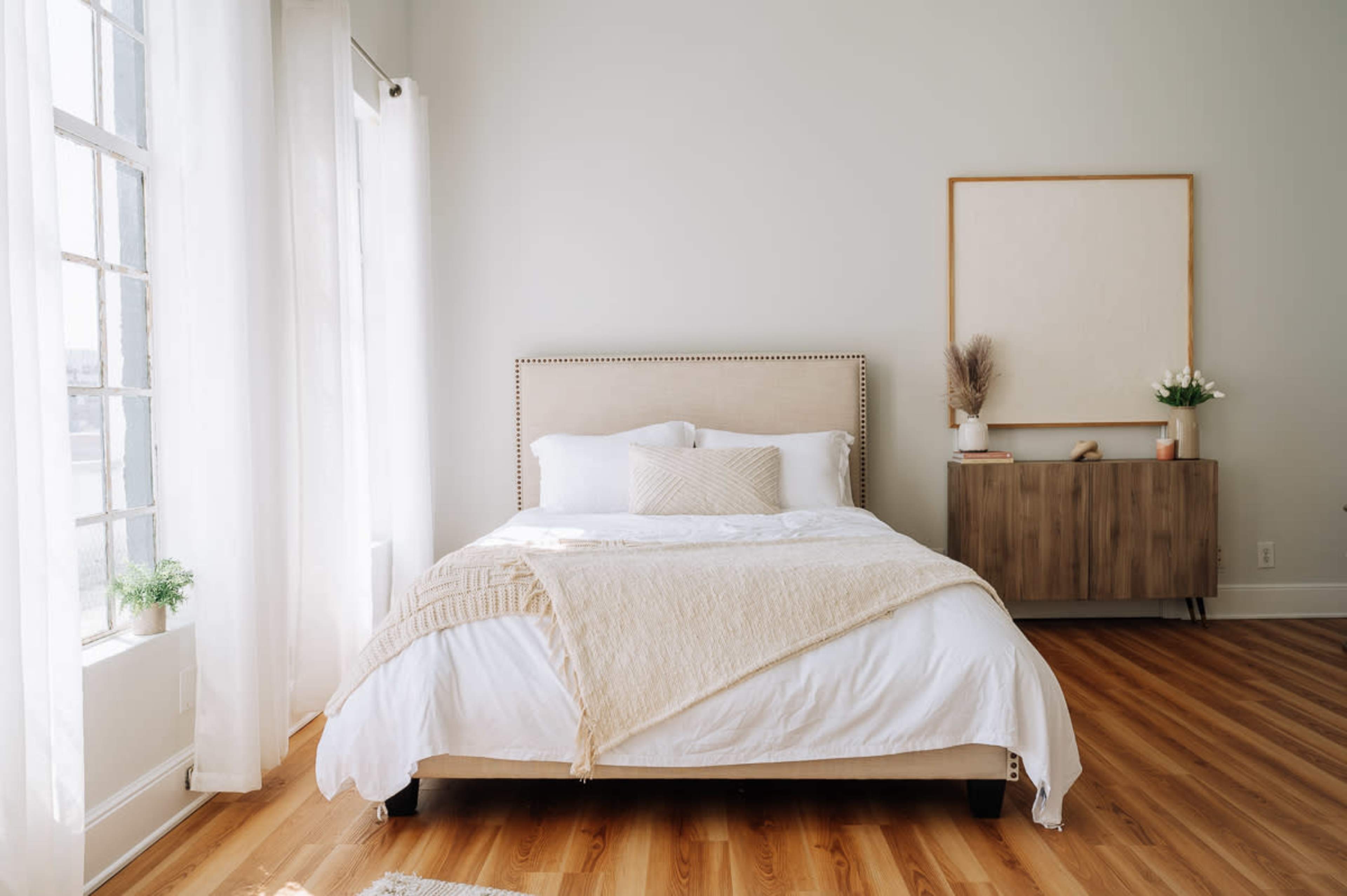 The image shows a bright bedroom featuring a bed with white bedding, a light-colored headboard, and a wooden dresser beside a large window with sheer curtains.