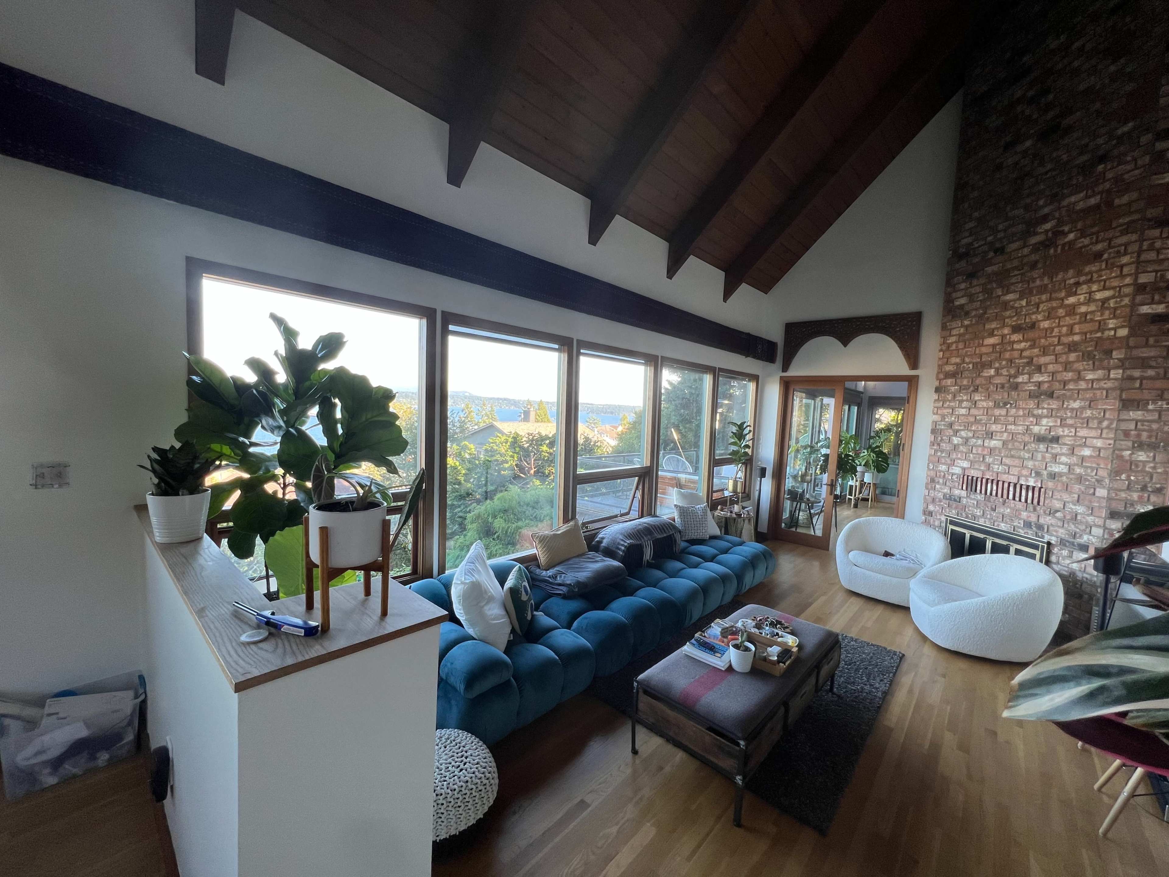 A spacious living room features a blue sofa, a coffee table, large windows with views of nature, and a high wooden ceiling.