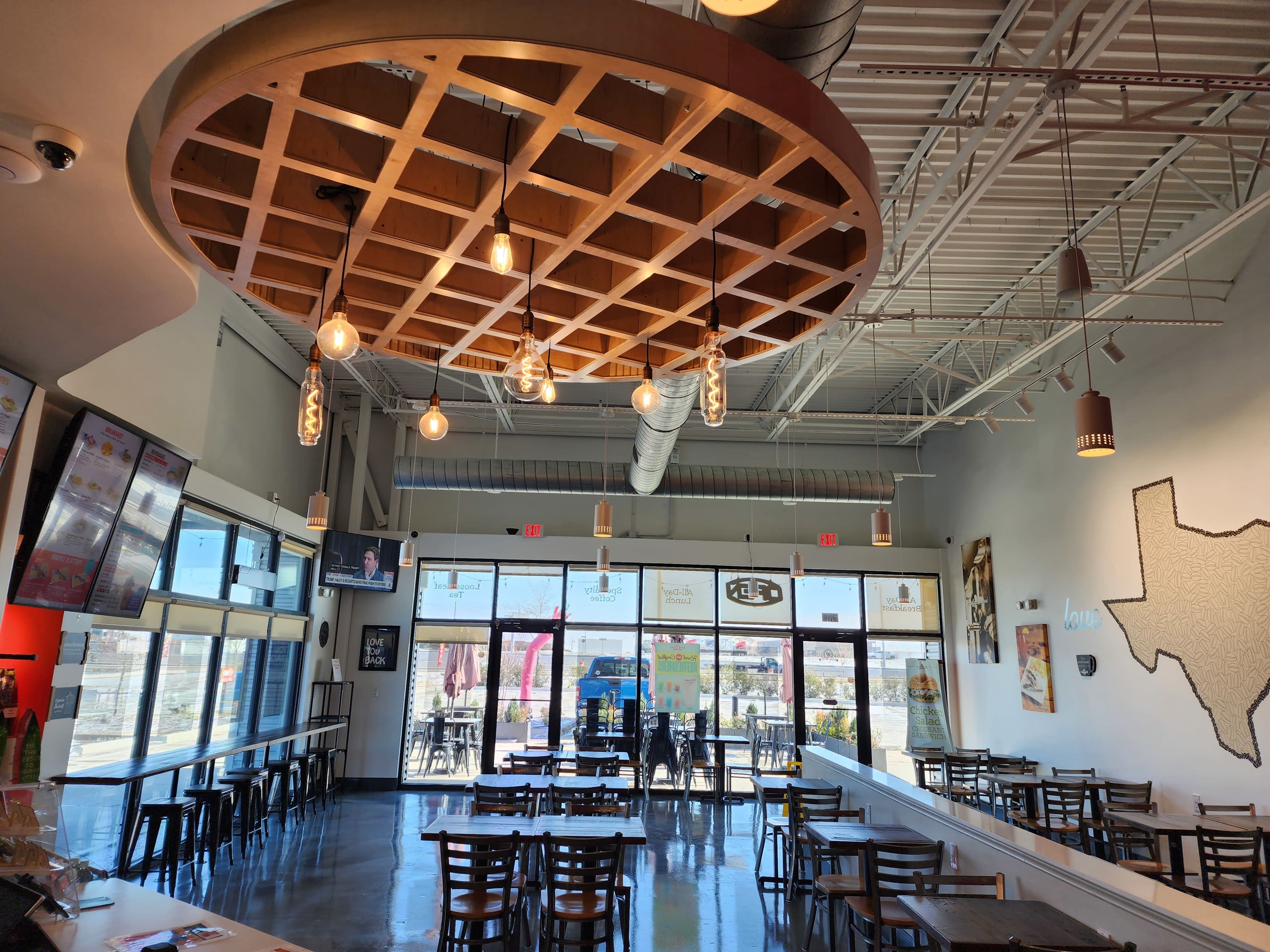 A modern café interior with a grid-patterned wooden ceiling feature, pendant lights, and rows of tables arranged for customers.