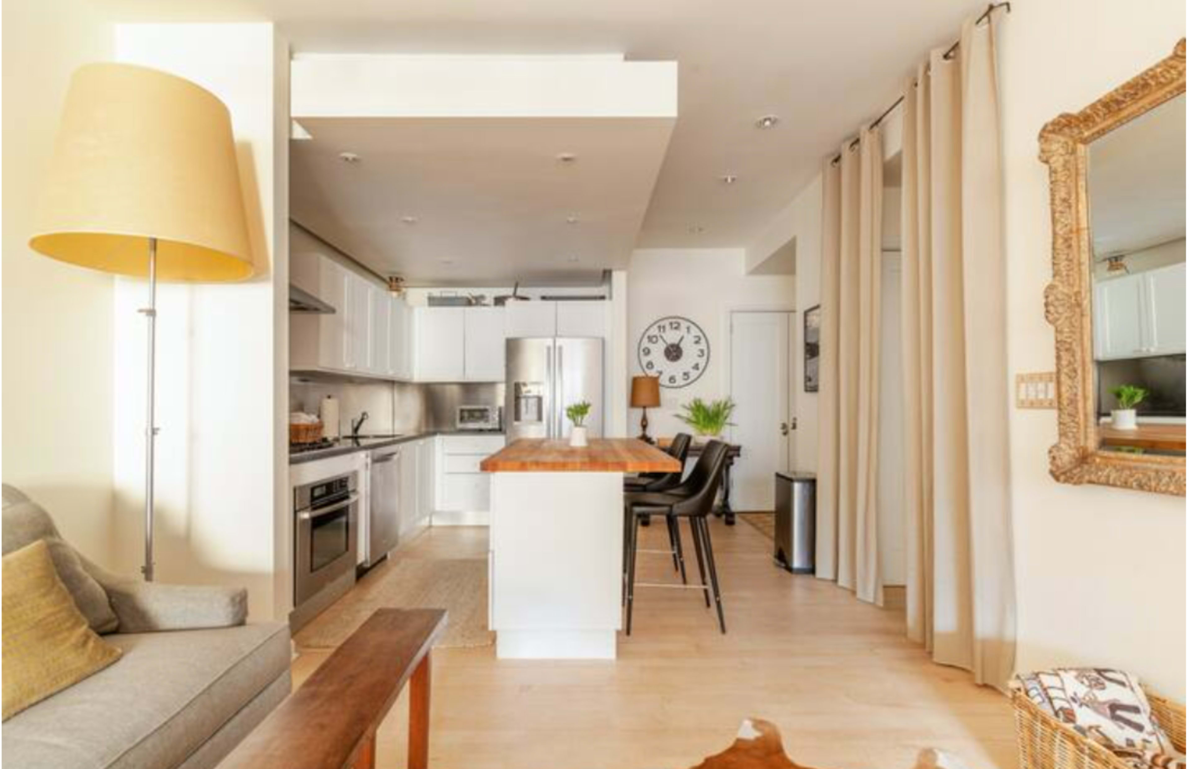 A modern kitchen and living area with a wooden island, white cabinetry, and a dining table set against a backdrop of light-colored walls and a large mirror.