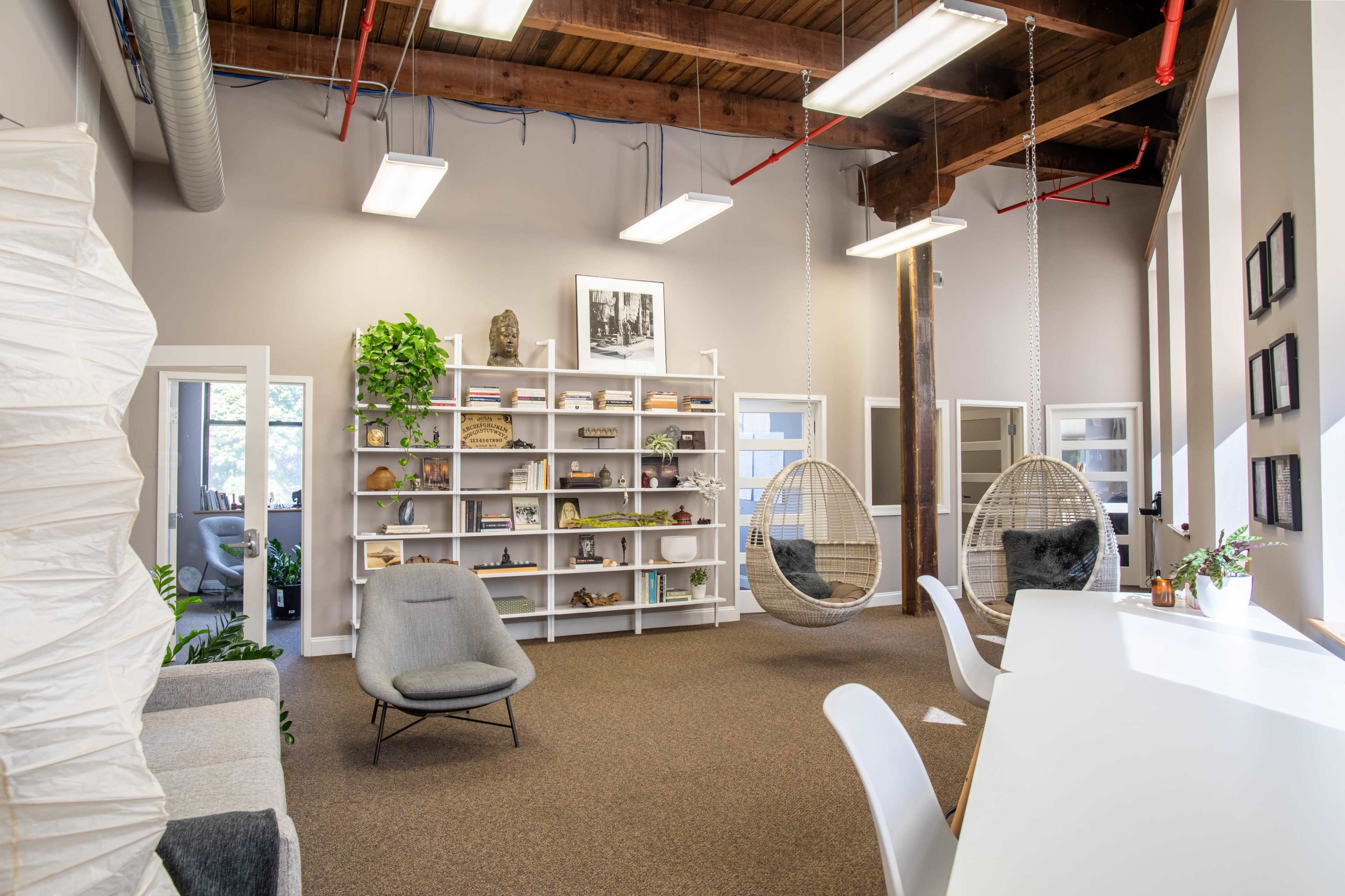 The image shows a modern office space featuring a seating area with hanging chairs, a bookshelf filled with decorative items, and large windows that provide natural light.