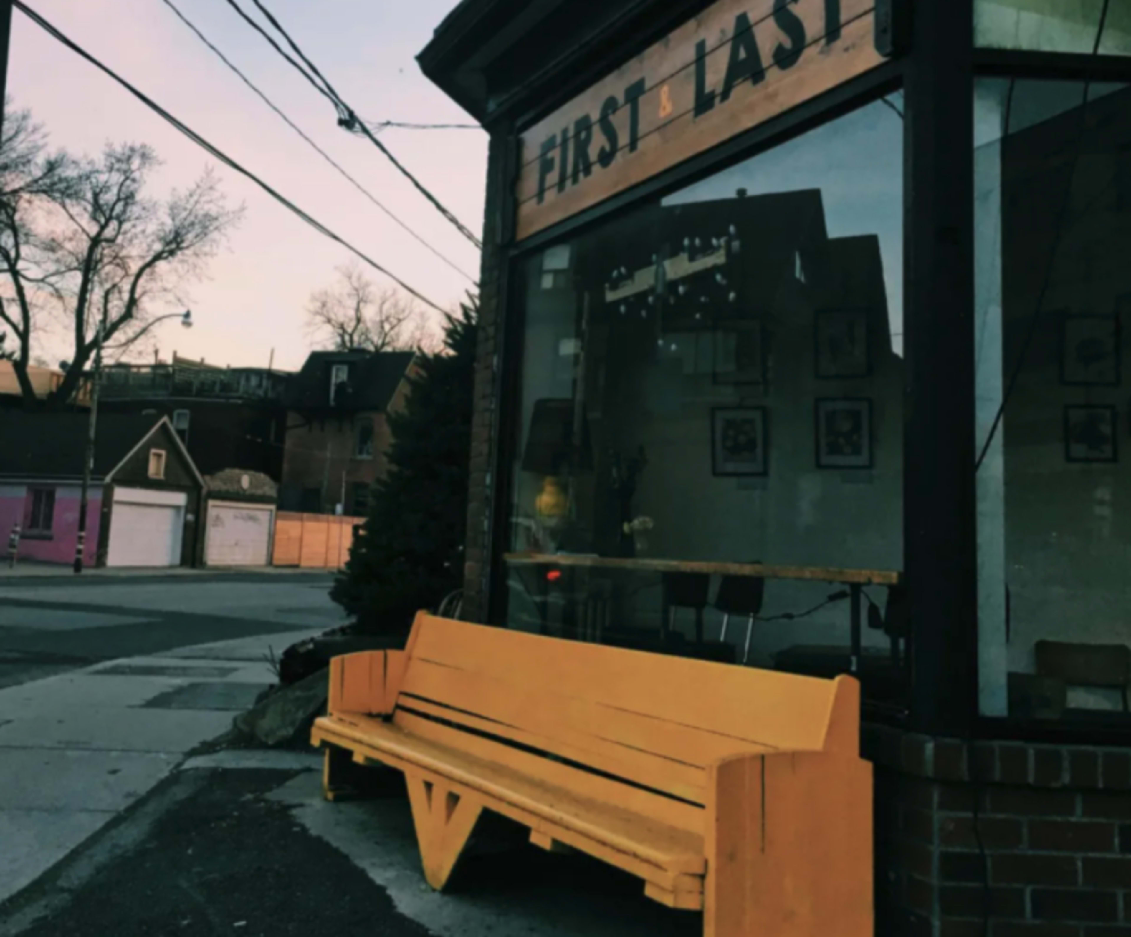 A bright yellow bench is positioned outside a building with a sign that reads "FIRST & LAST," reflecting the evening light.