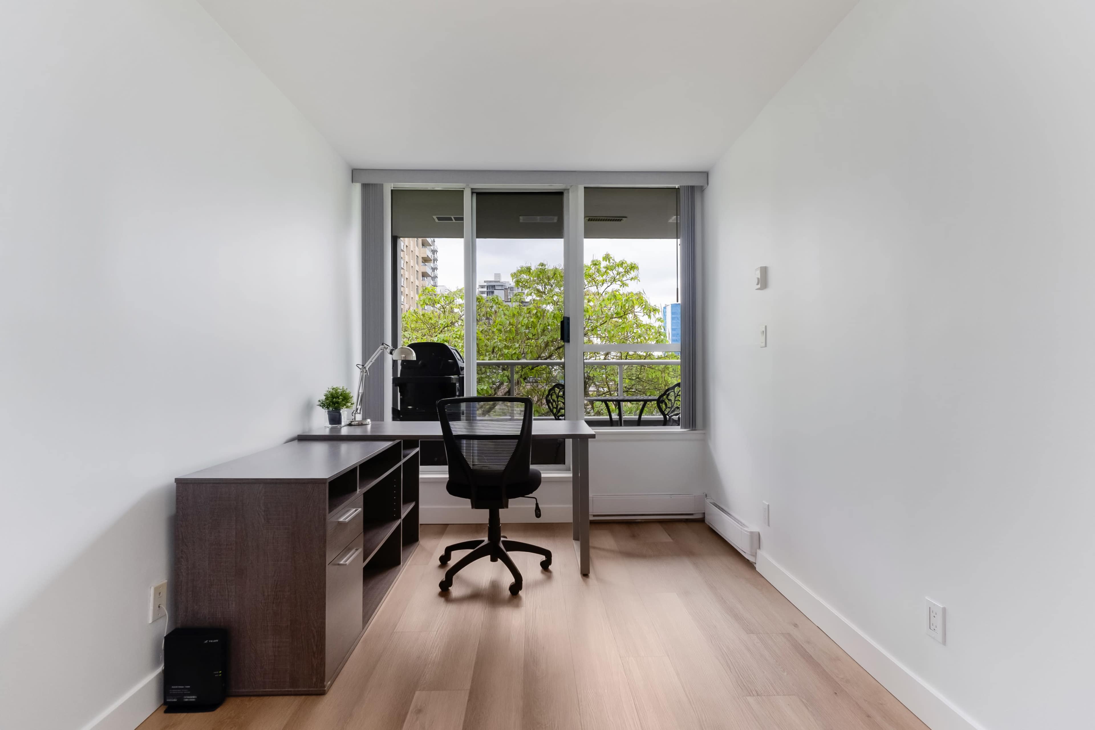 A minimalist workspace features a wooden desk and an office chair beside a large window with a view of greenery.
