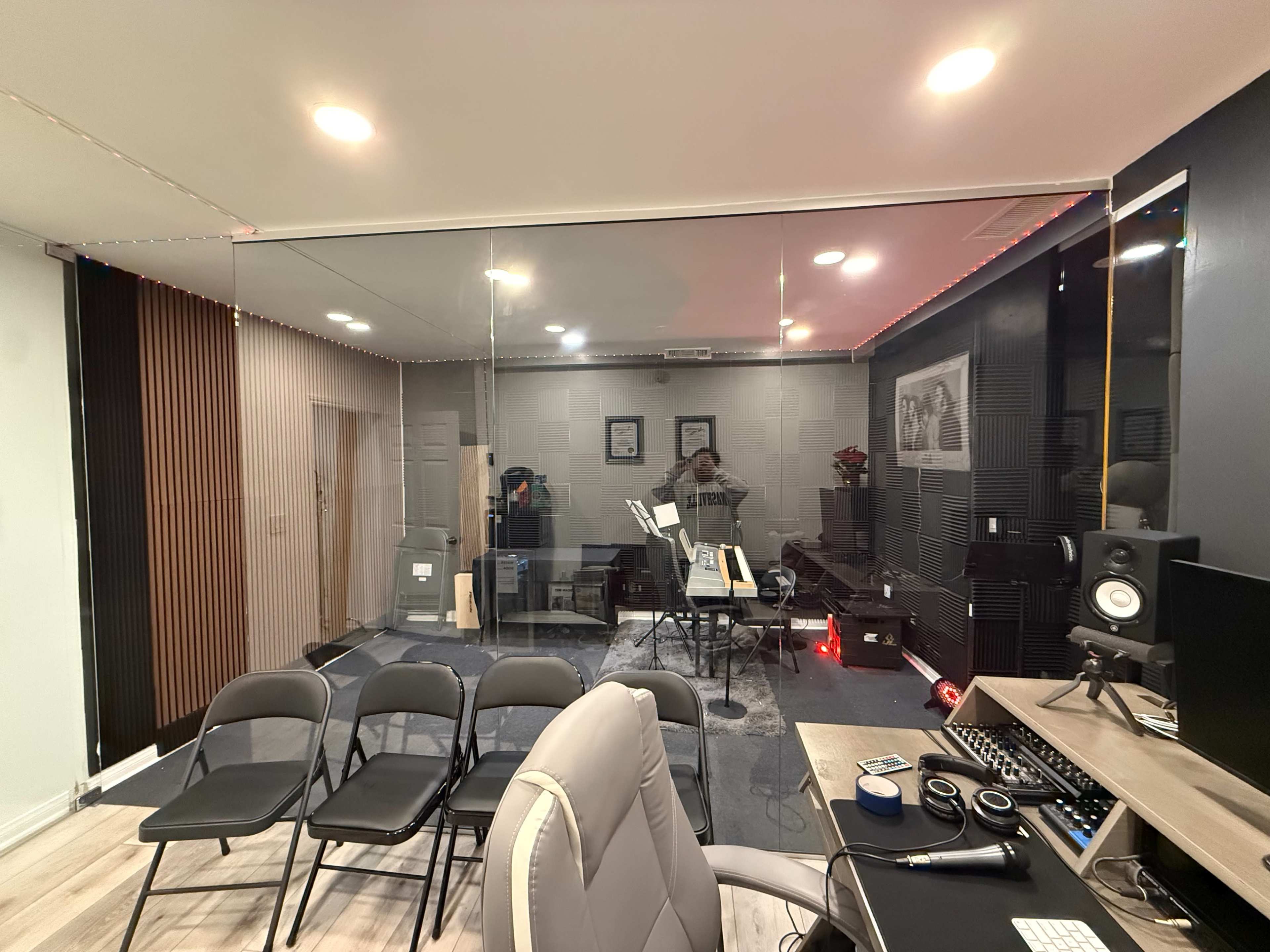 The image shows a well-lit music studio with a glass wall, featuring audio equipment and seating arrangements.