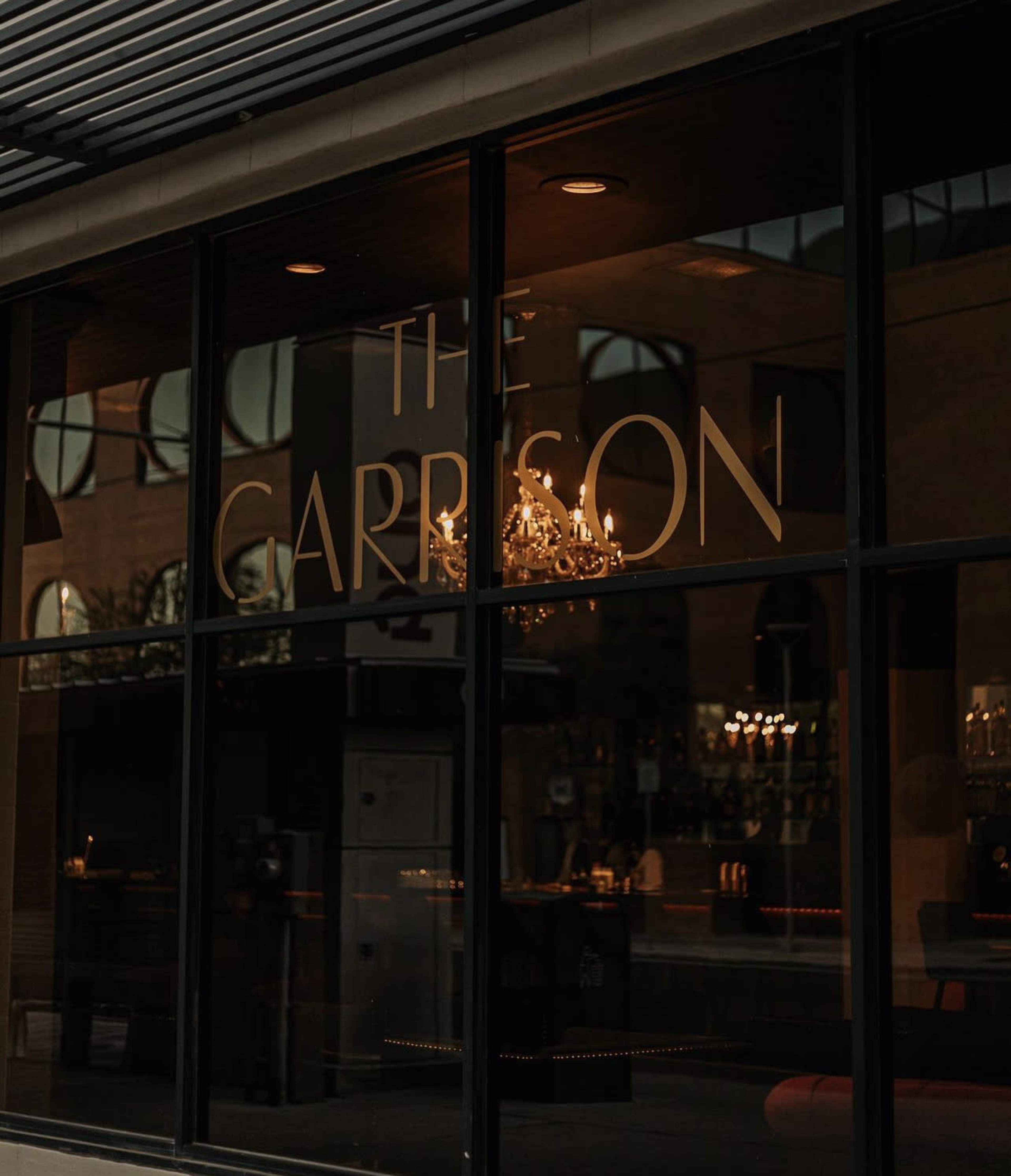 The entrance of a building with the sign "The Garrison" reflected in a large glass window, featuring a chandelier inside.
