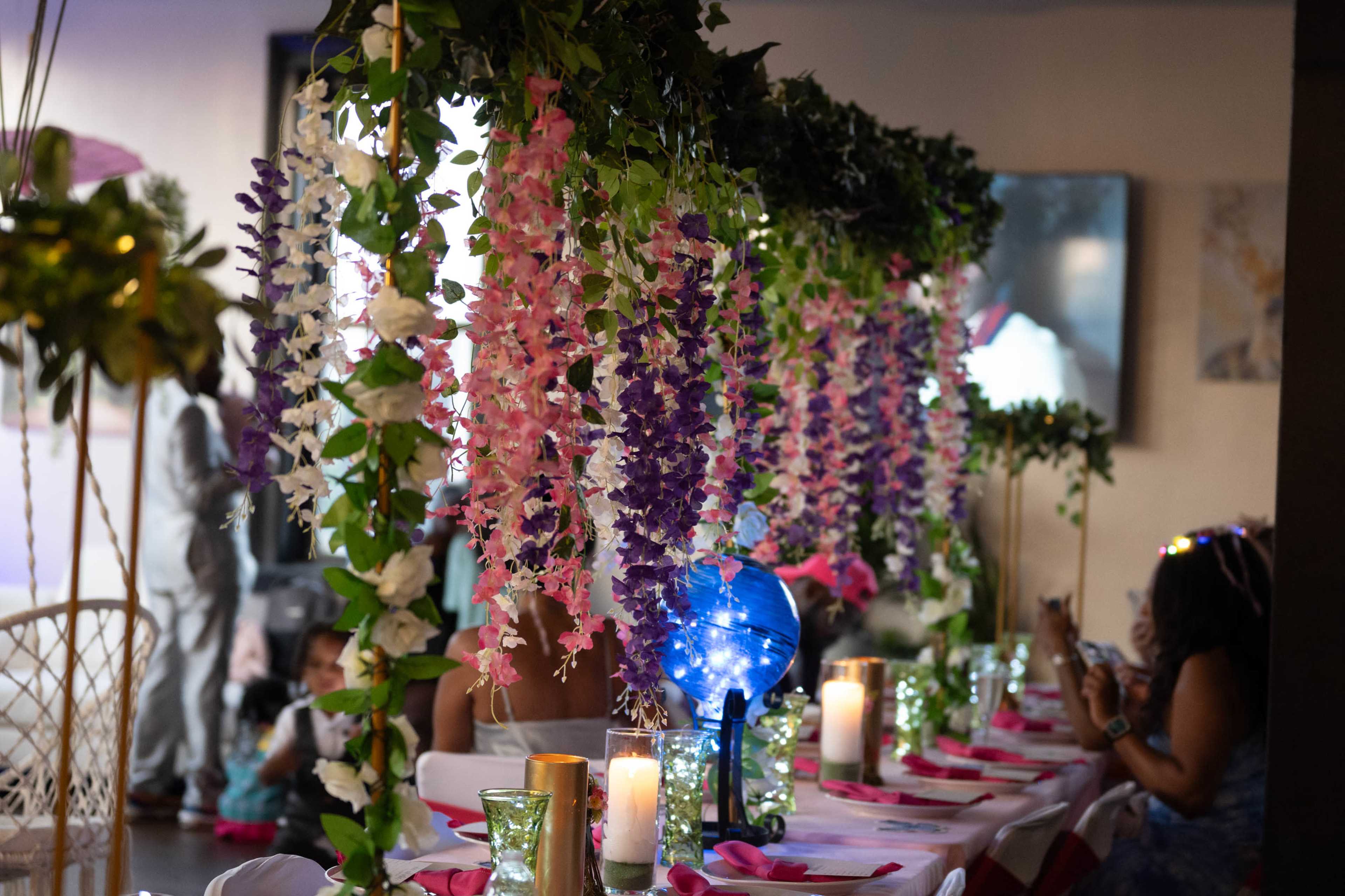 A decorated dining area features rows of flowers hanging from a canopy above tables set with candles and colorful tableware.