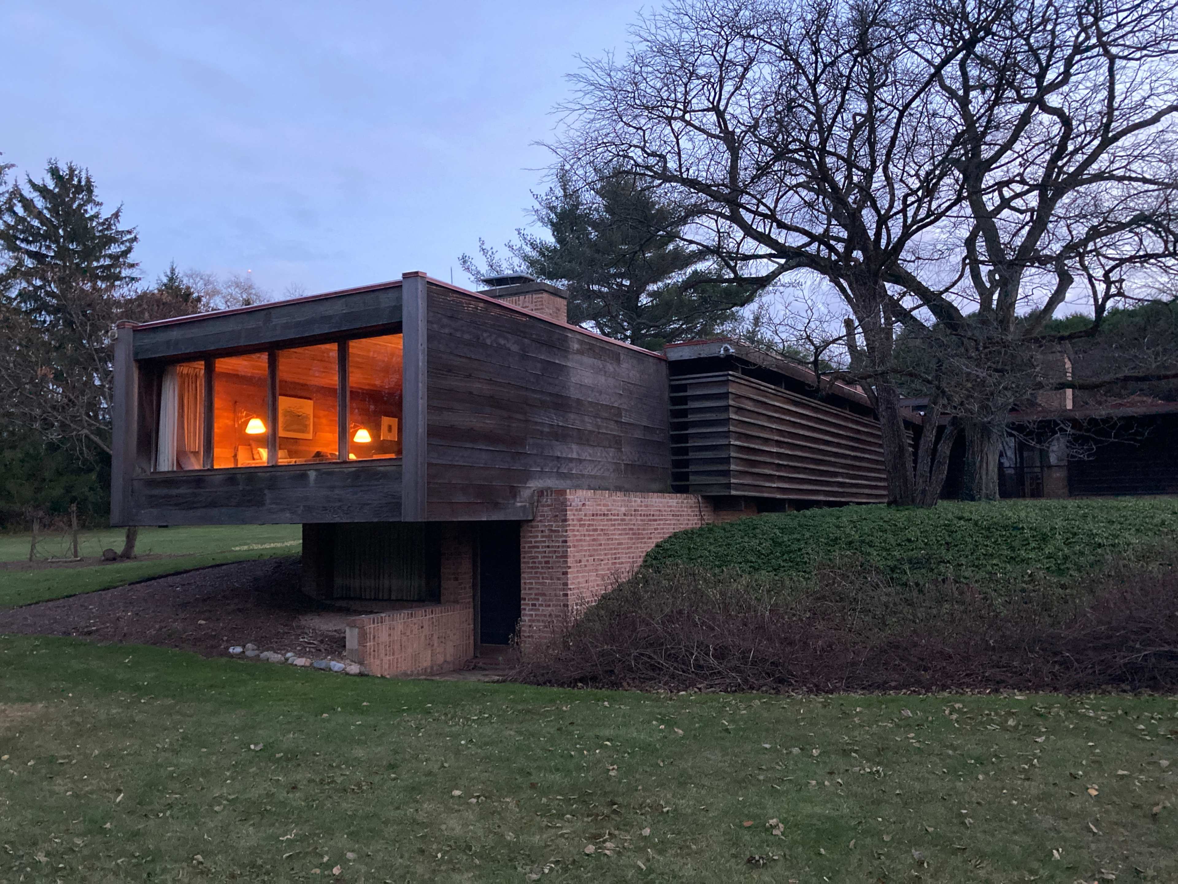 A modern wooden house with large windows extends over a brick foundation, surrounded by trees and grassy land.