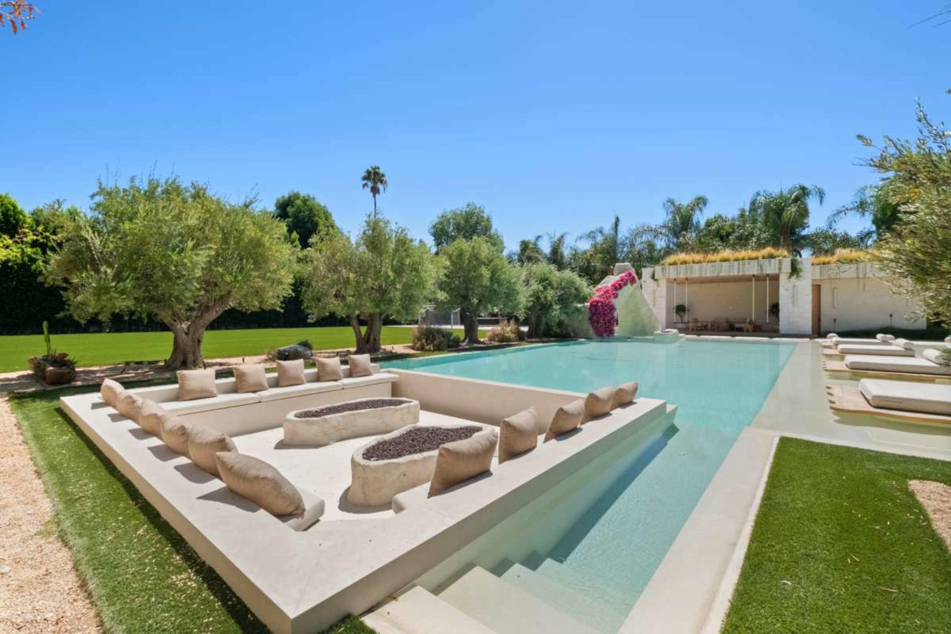 The image shows a modern outdoor swimming pool area surrounded by green landscaping, featuring sun loungers and seating arrangements made of light-colored materials.