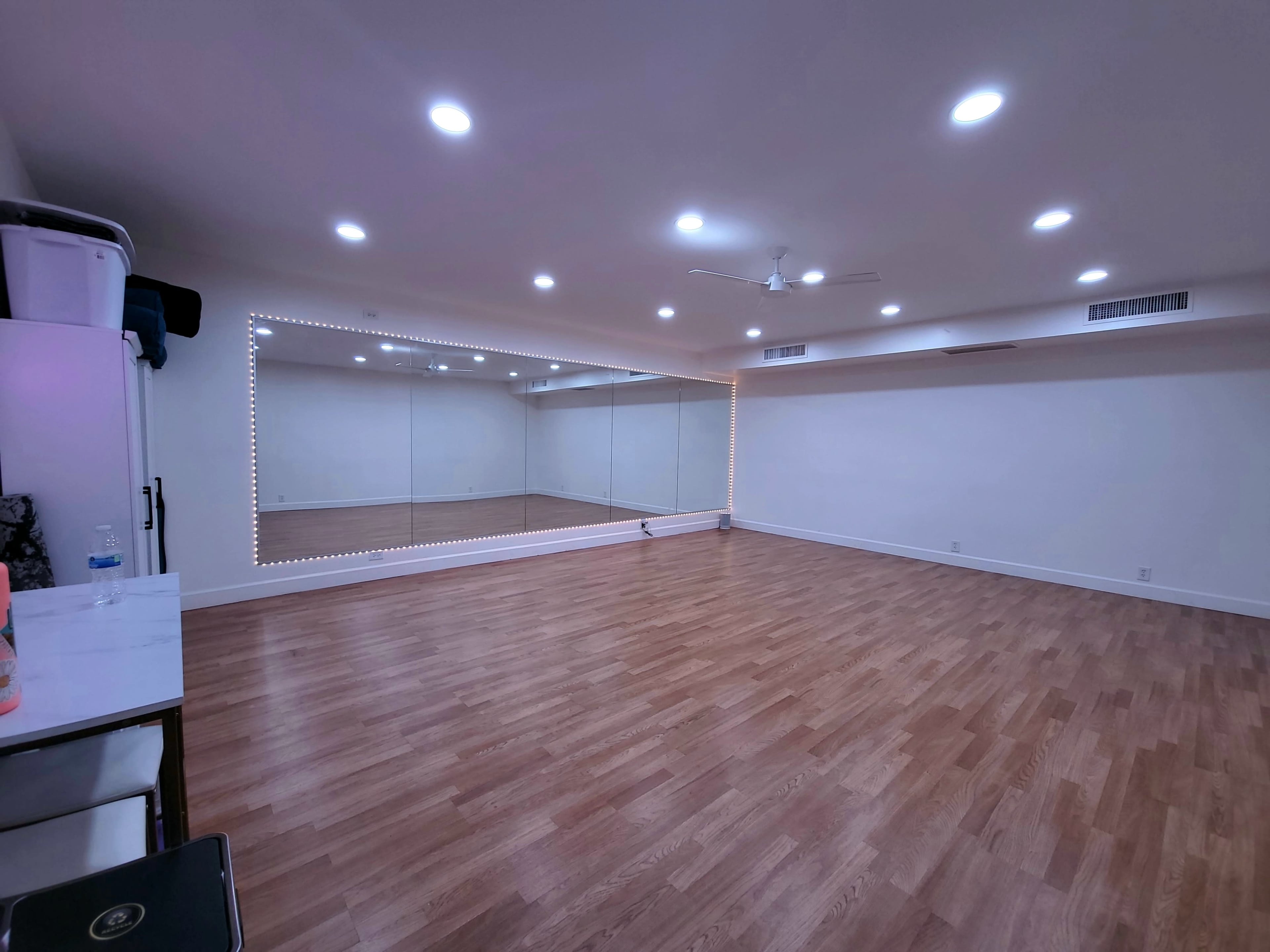 A spacious, well-lit room with wooden flooring and a large mirror framed by lights on one wall.