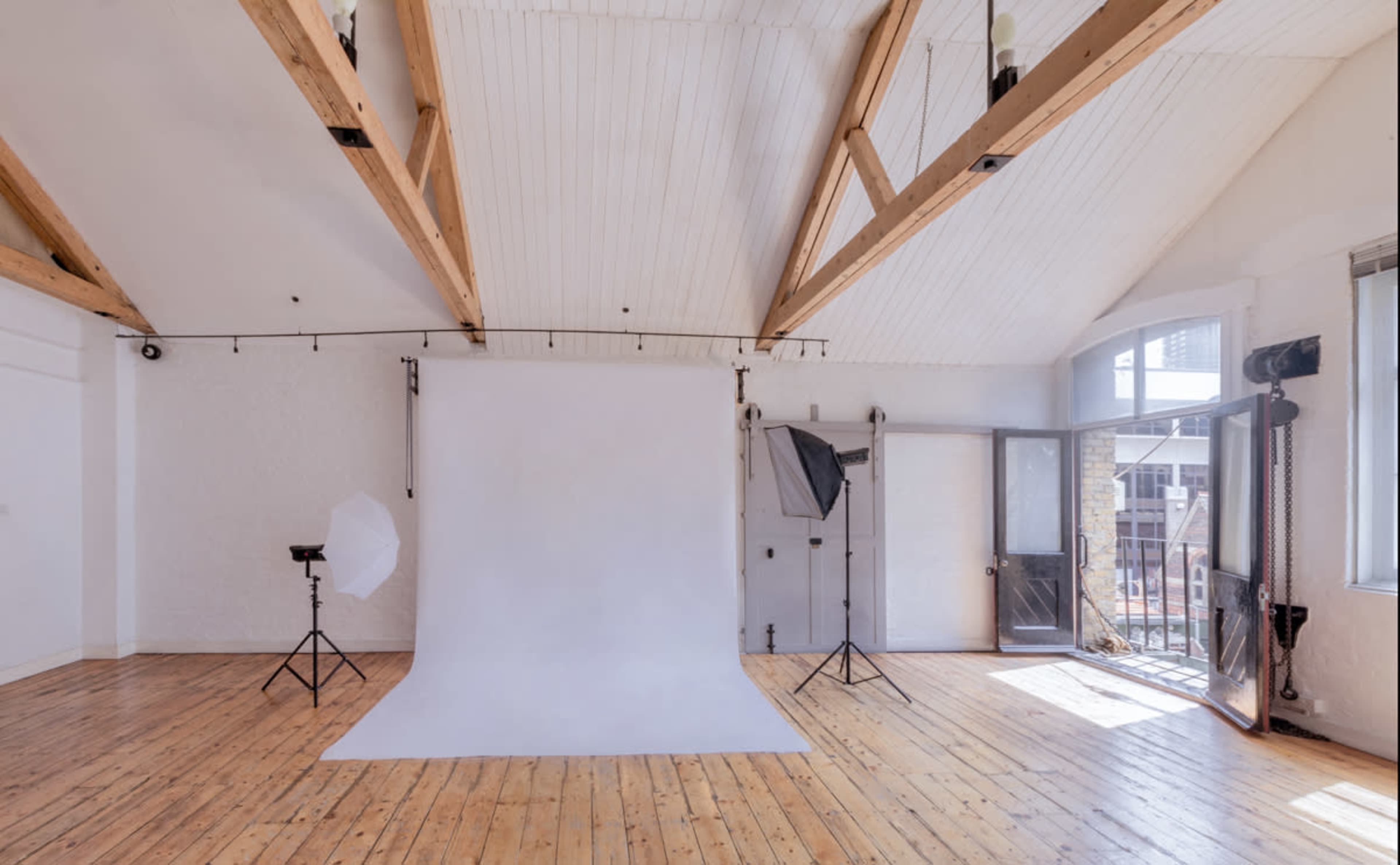 A photography studio with a white backdrop, wooden floors, and large windows, complemented by studio lighting equipment.