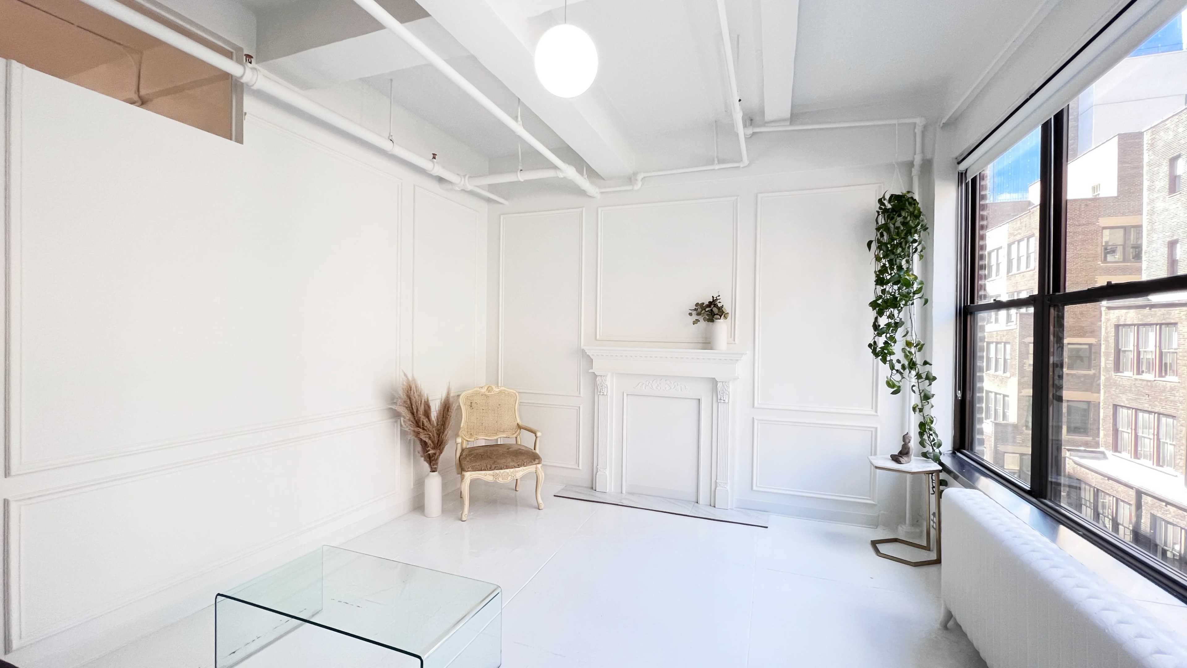 A bright, minimalist room with white walls, a decorative fireplace, a single chair, and large windows letting in natural light.