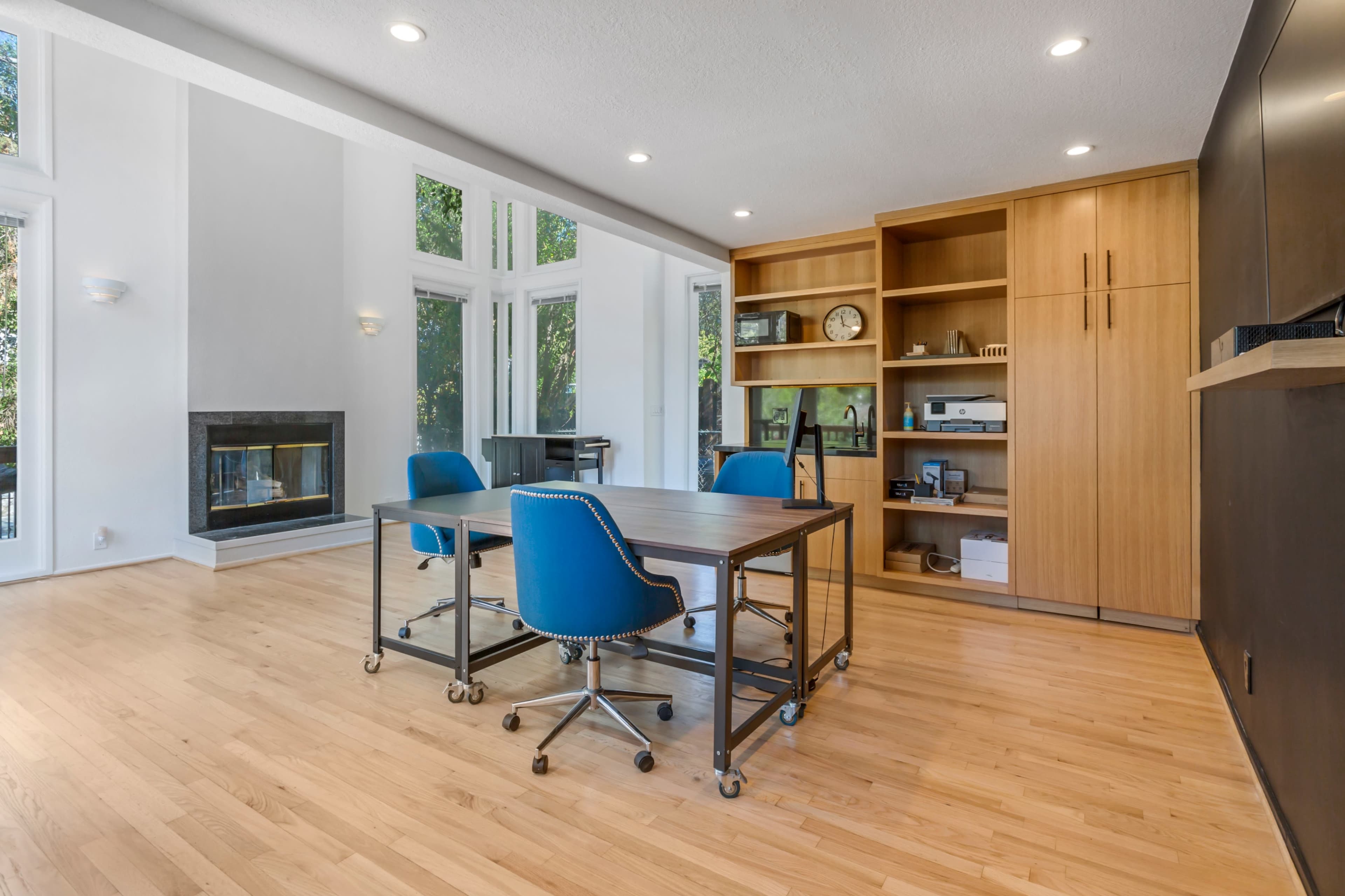 A modern workspace features a wooden table with chairs on wheels and built-in shelving, surrounded by large windows that bring in natural light.