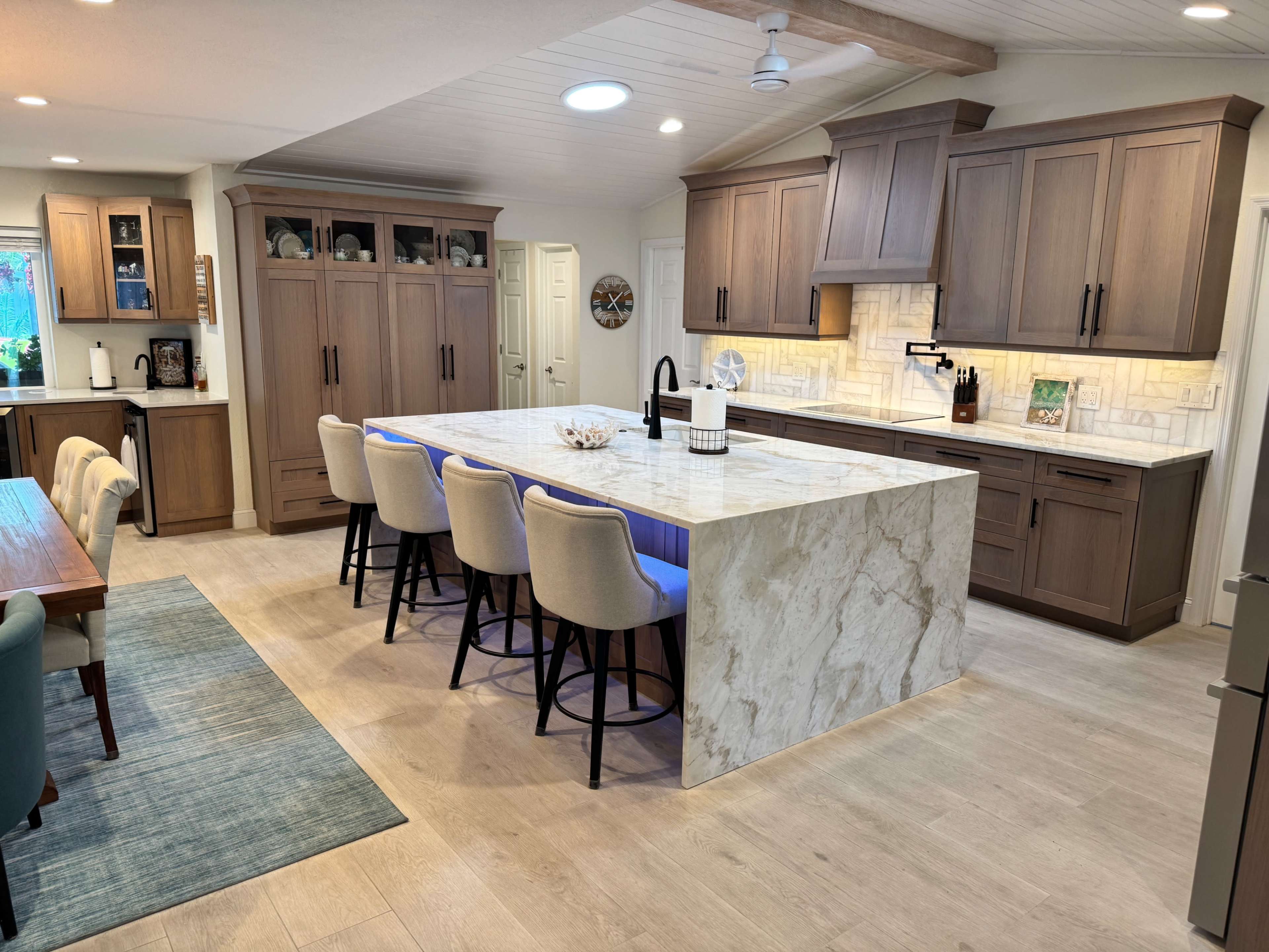 The image depicts a modern kitchen with an island featuring seating, wooden cabinetry, and a light-colored stone countertop.