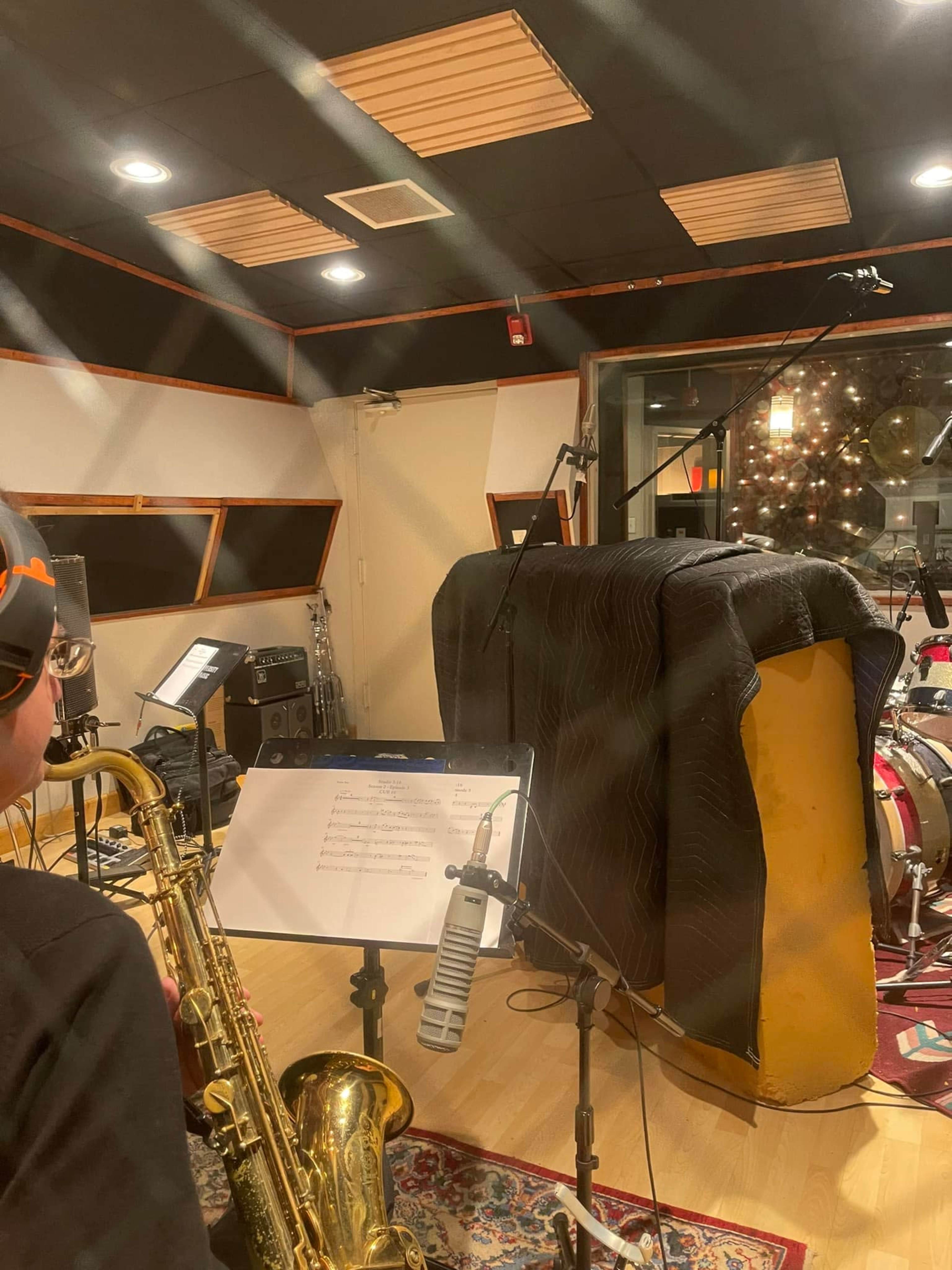 A musician plays a saxophone in a recording studio, with a microphone set up in front of sheet music and various instruments in the background.