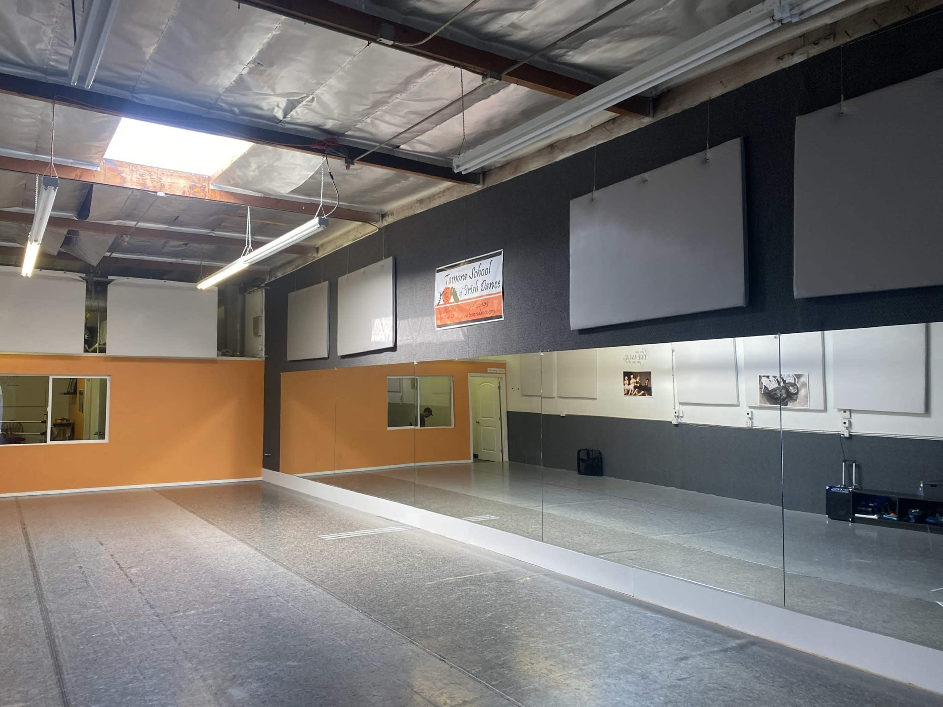 A dance studio with a large mirror on one wall, orange and gray walls, and overhead fluorescent lighting.