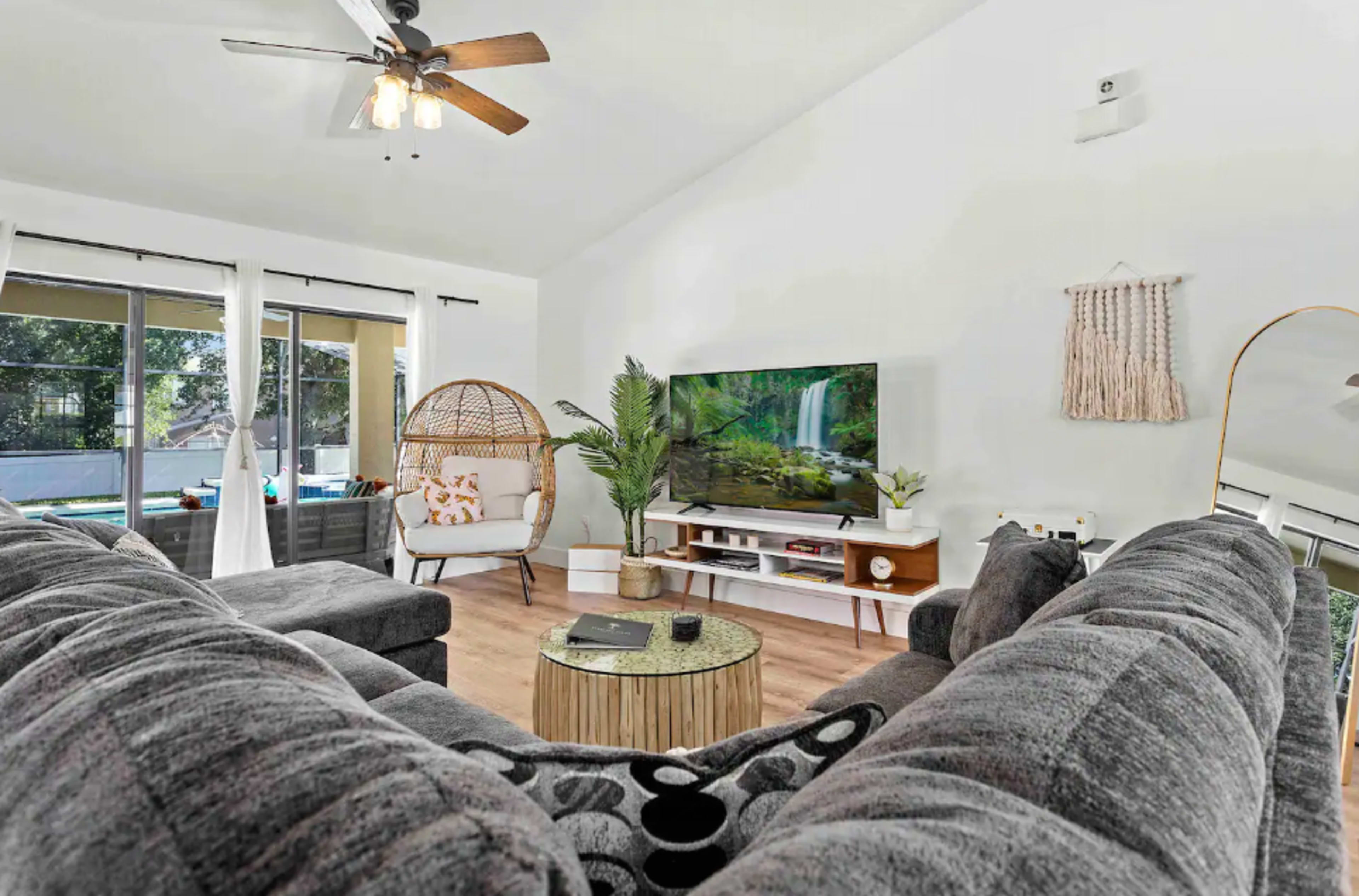 The image shows a modern living room featuring a large sectional sofa, a television mounted on the wall, and a hanging chair near a window.