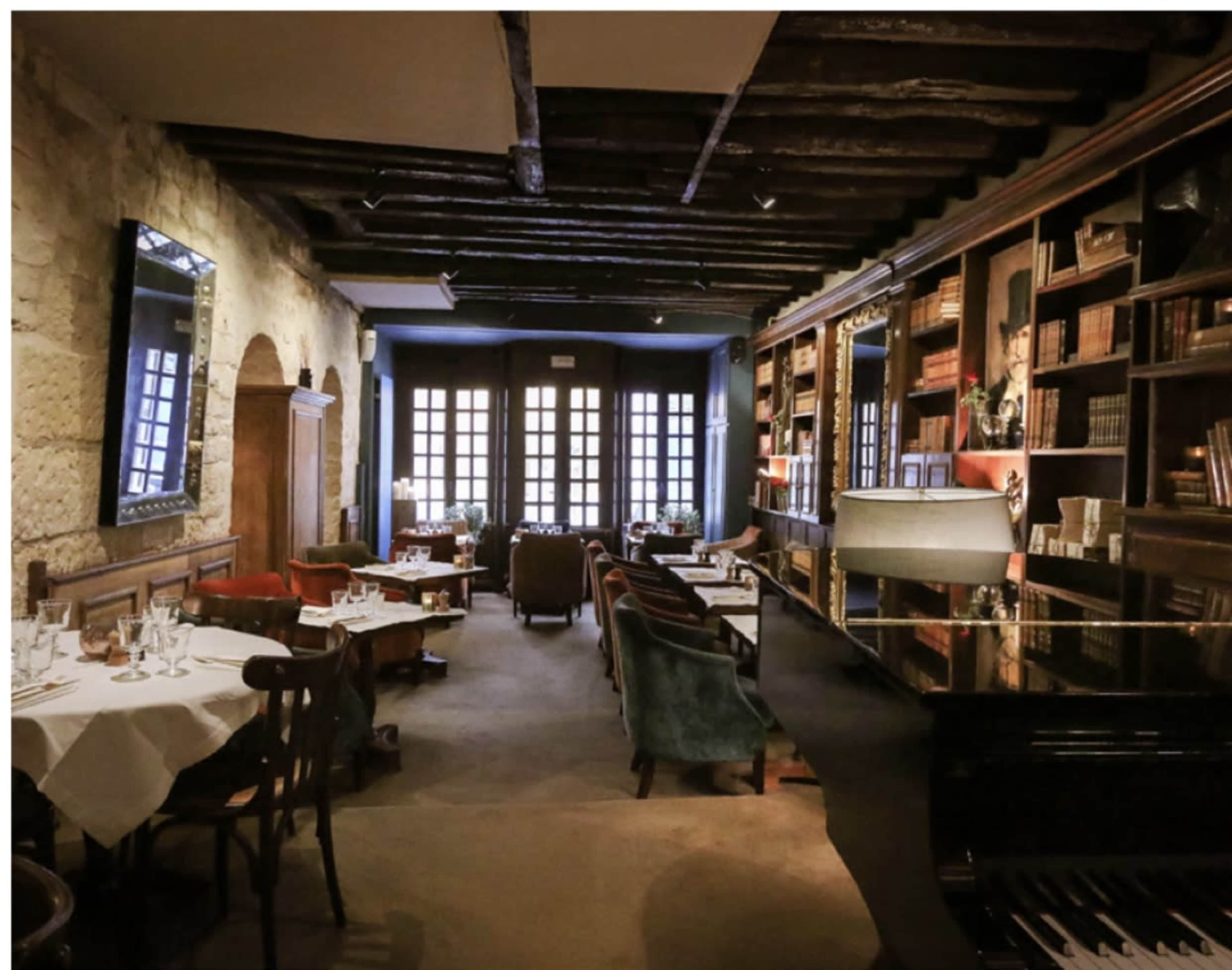 A dimly lit restaurant with wooden beams, tables set for dining, bookshelves lining the walls, and a piano in the corner.