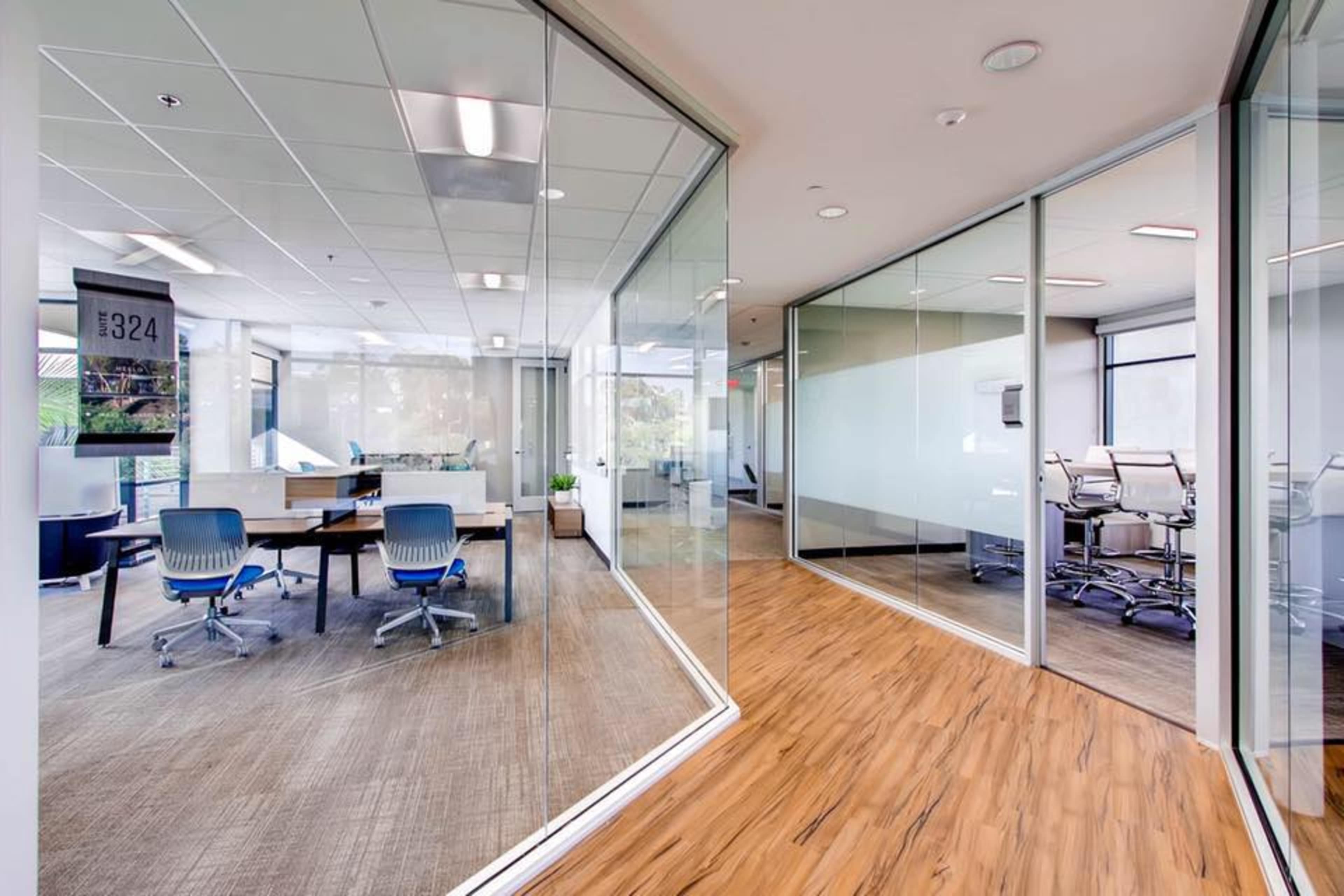 The image shows a modern office space featuring glass-walled meeting rooms, a communal area with desks, and wooden flooring.