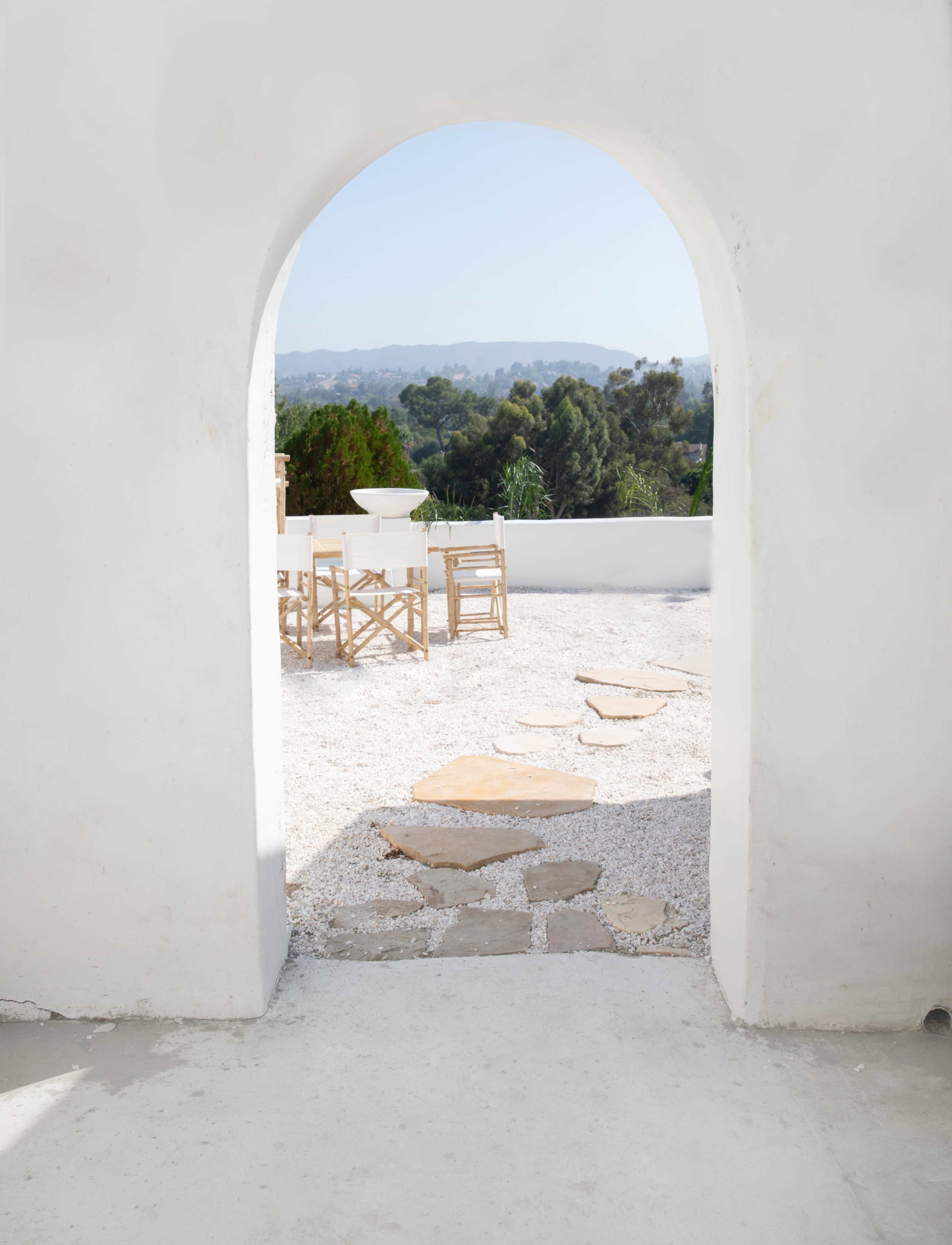 A white archway frames a pathway leading to an outdoor area with wooden furniture and a view of distant trees and mountains.