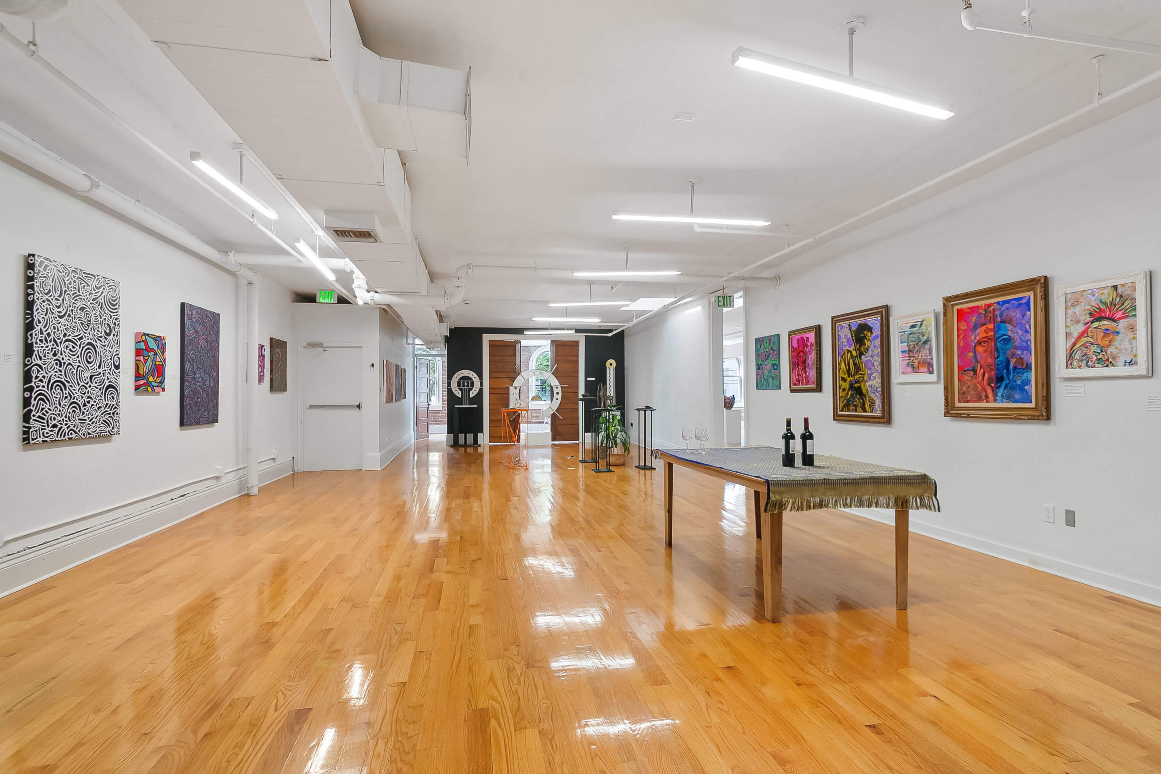 The image shows a gallery space with polished wooden floors, white walls adorned with colorful artwork, and a central table set with three decorative items.