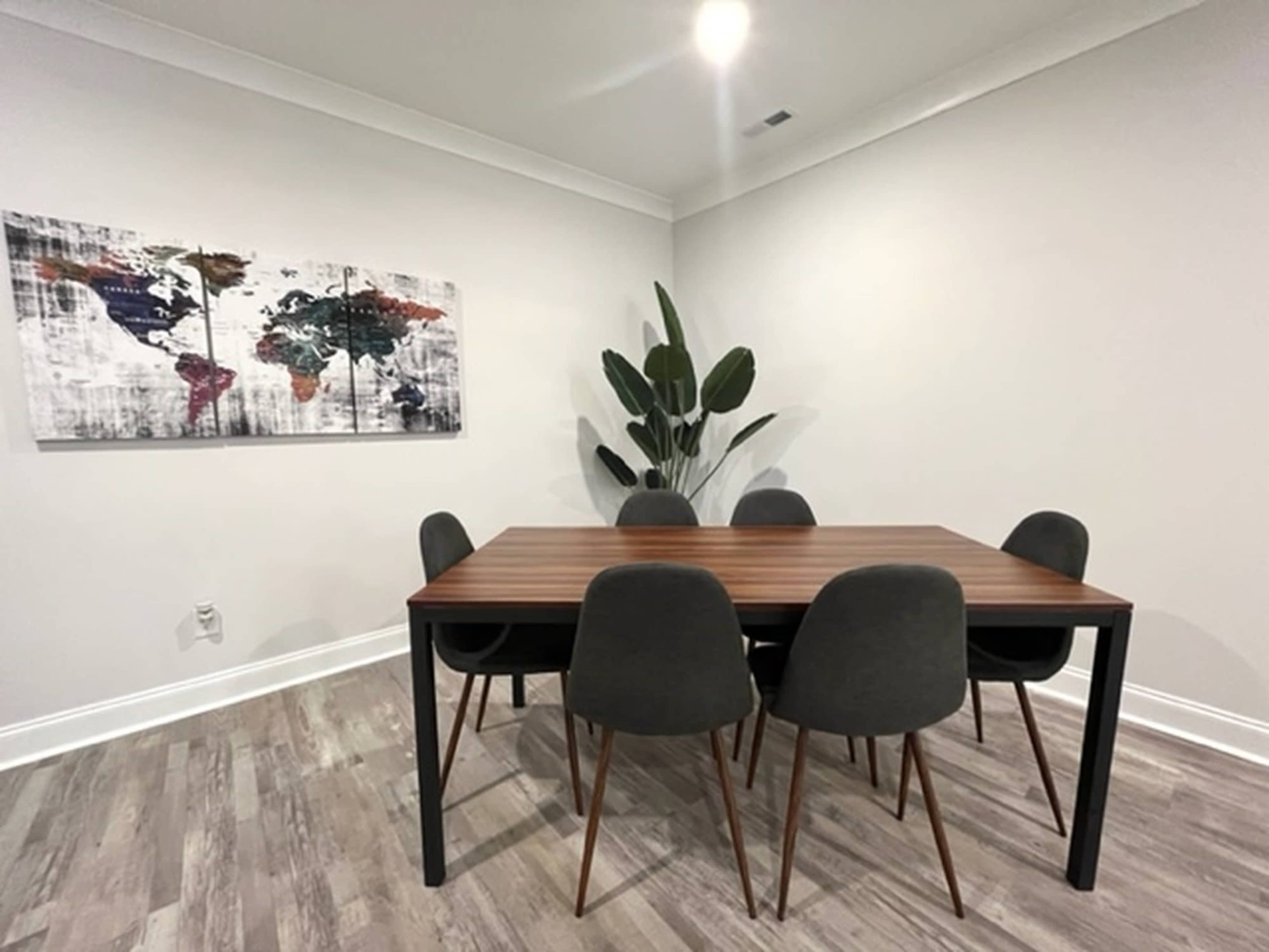 A wooden dining table surrounded by six chairs is positioned in a light-colored room featuring a world map artwork on the wall and a tall plant in the corner.