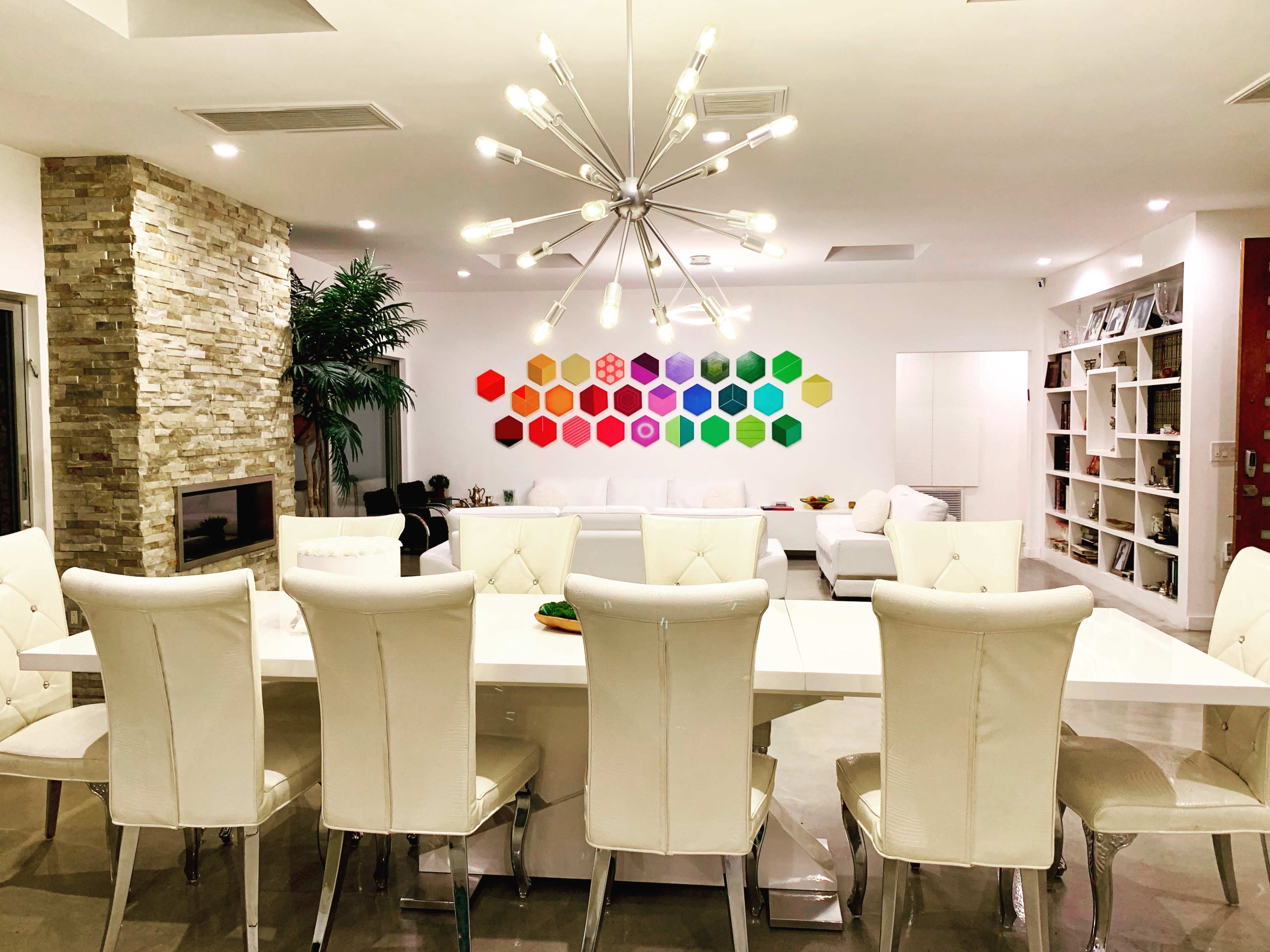 The image shows a modern living space featuring a long white dining table with eight white chairs, a colorful hexagon art installation on the wall, and a cozy seating area with a fireplace.