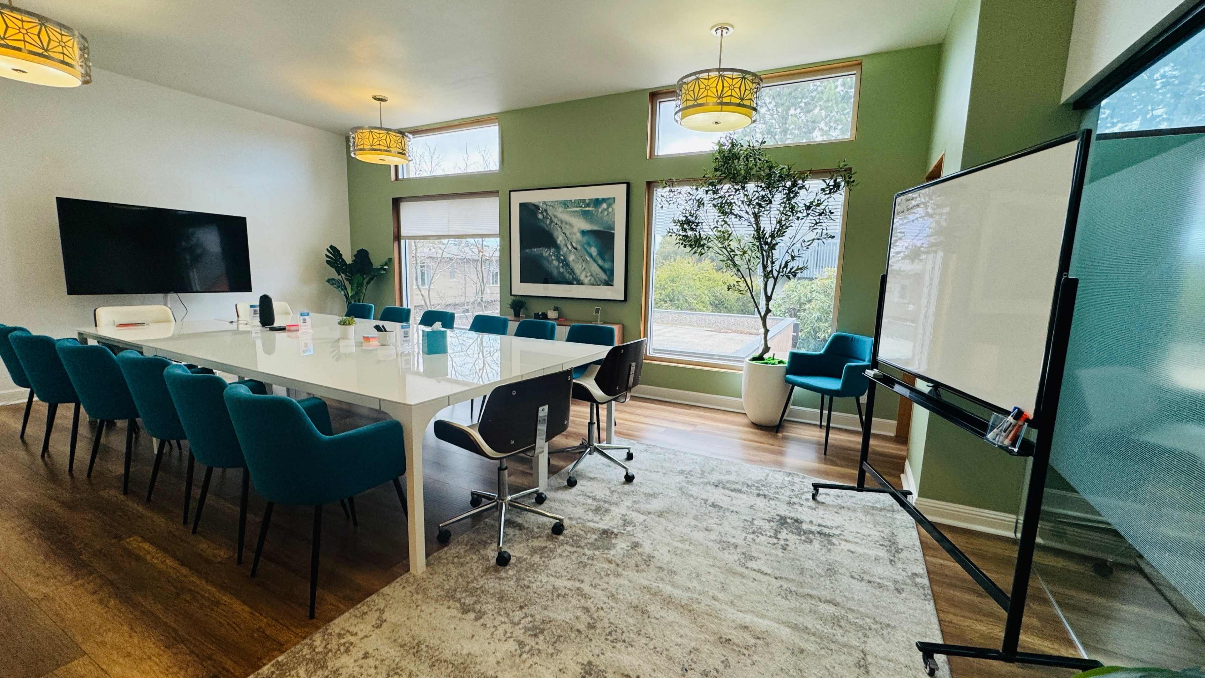 A modern conference room features a long white table surrounded by teal chairs, with large windows allowing natural light and a view of greenery outside.