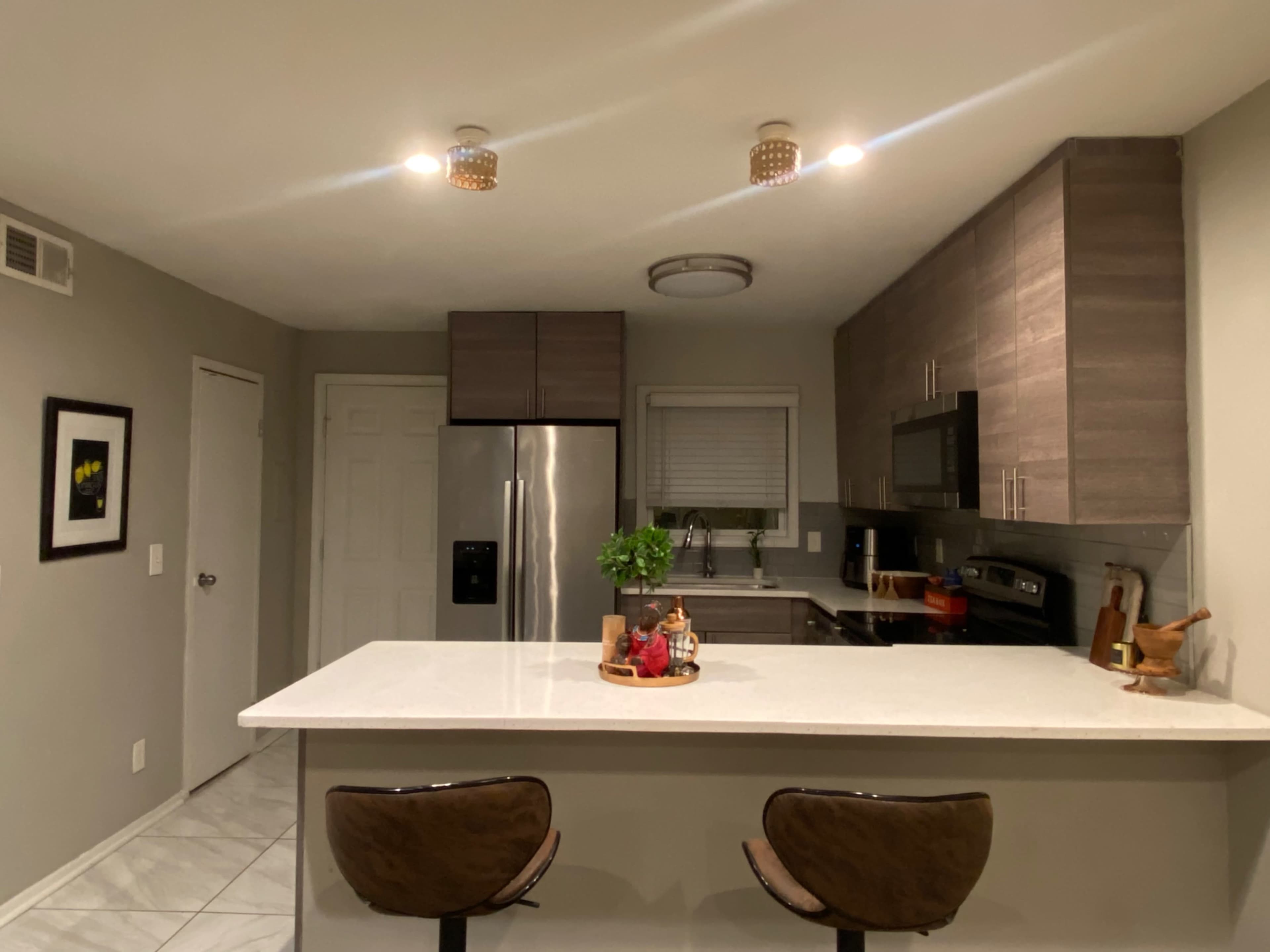 A modern kitchen featuring a central island with two bar stools, light cabinetry, stainless steel appliances, and recessed lighting.