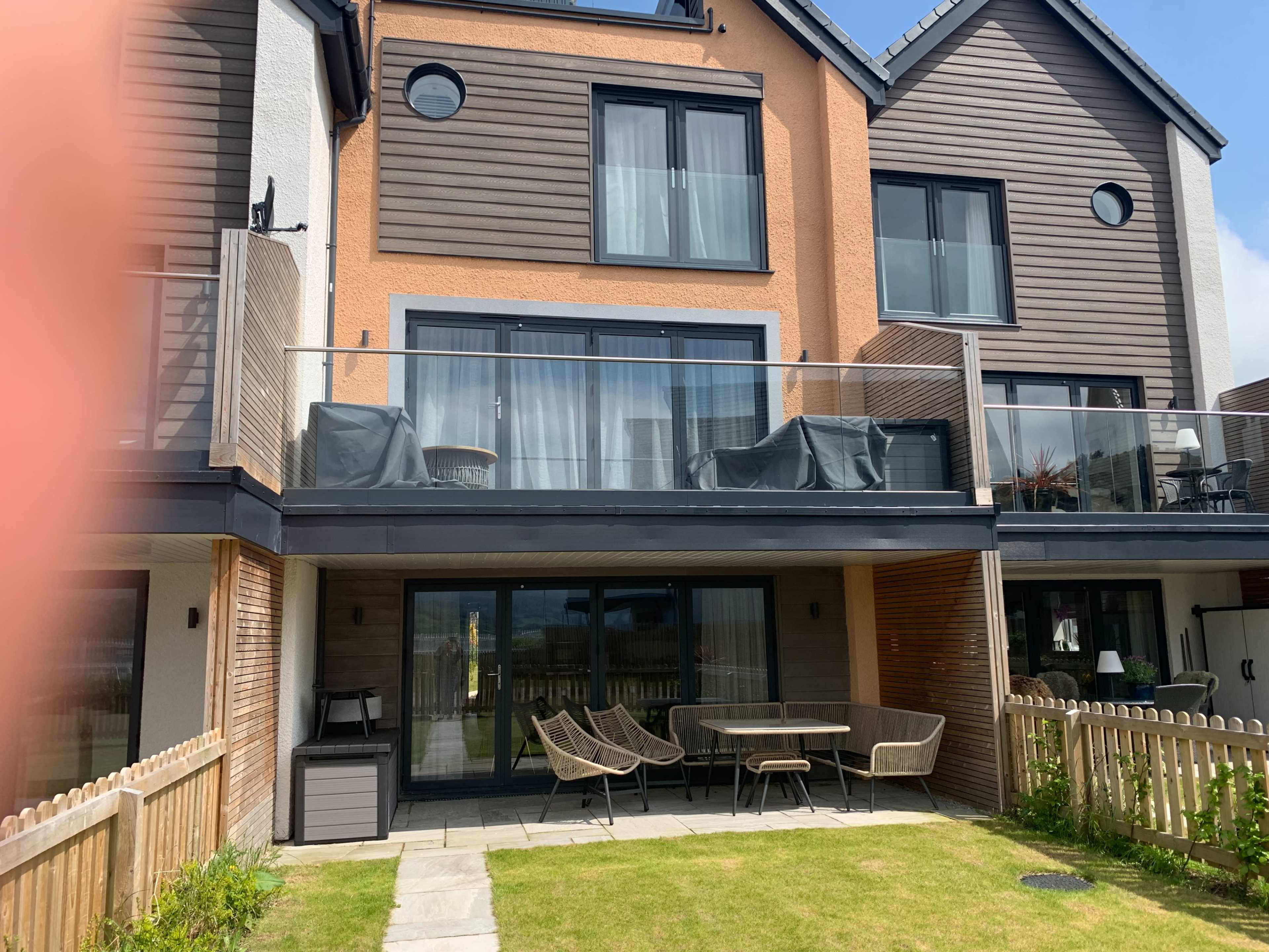 The image shows a modern two-story building with a terrace and patio furniture in a grassy yard.