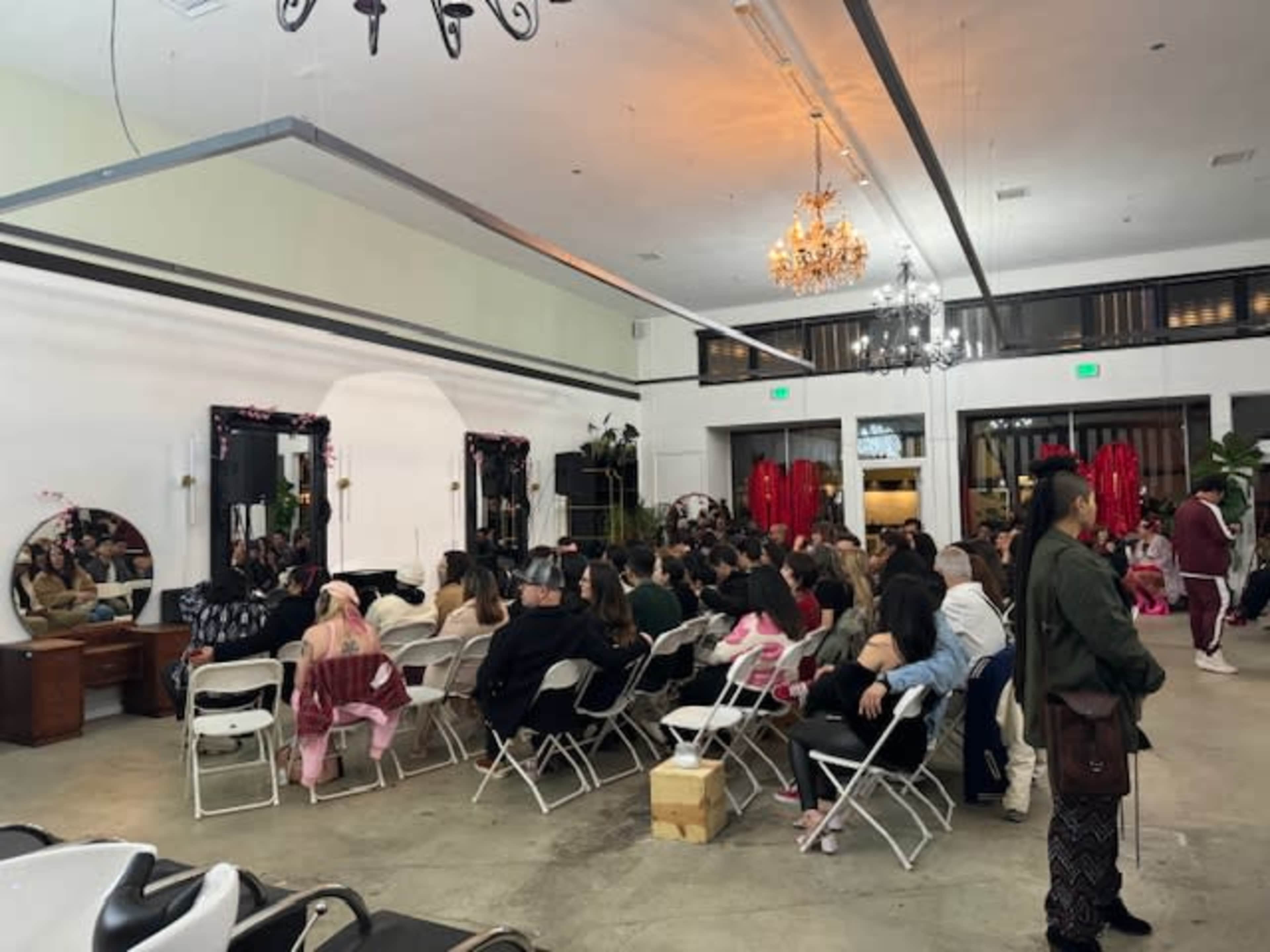 A large audience is seated in folding chairs in a spacious room featuring chandeliers and wall mirrors.