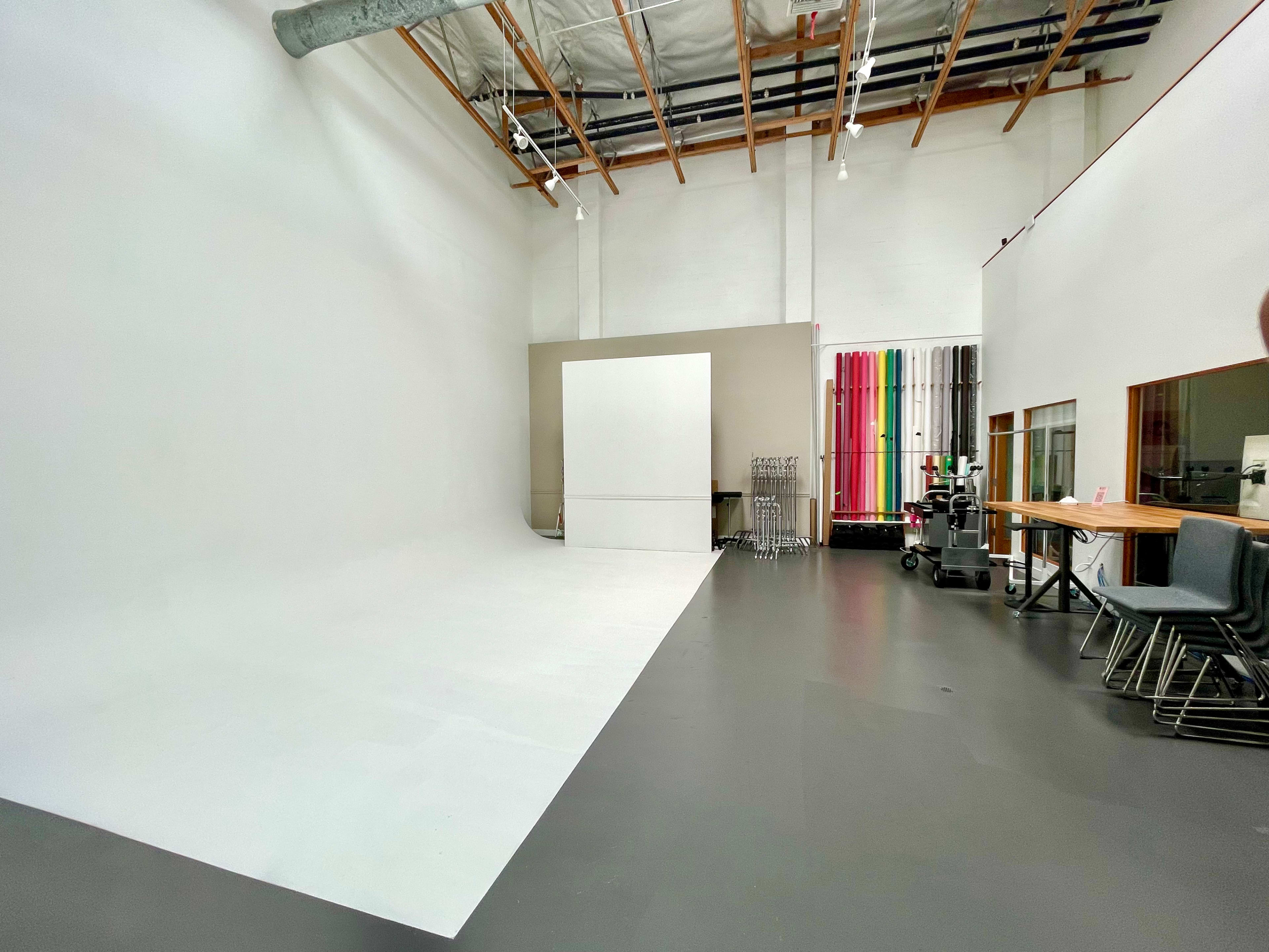 A spacious photography studio with a white backdrop, gray flooring, and colorful backdrops neatly arranged against the wall.