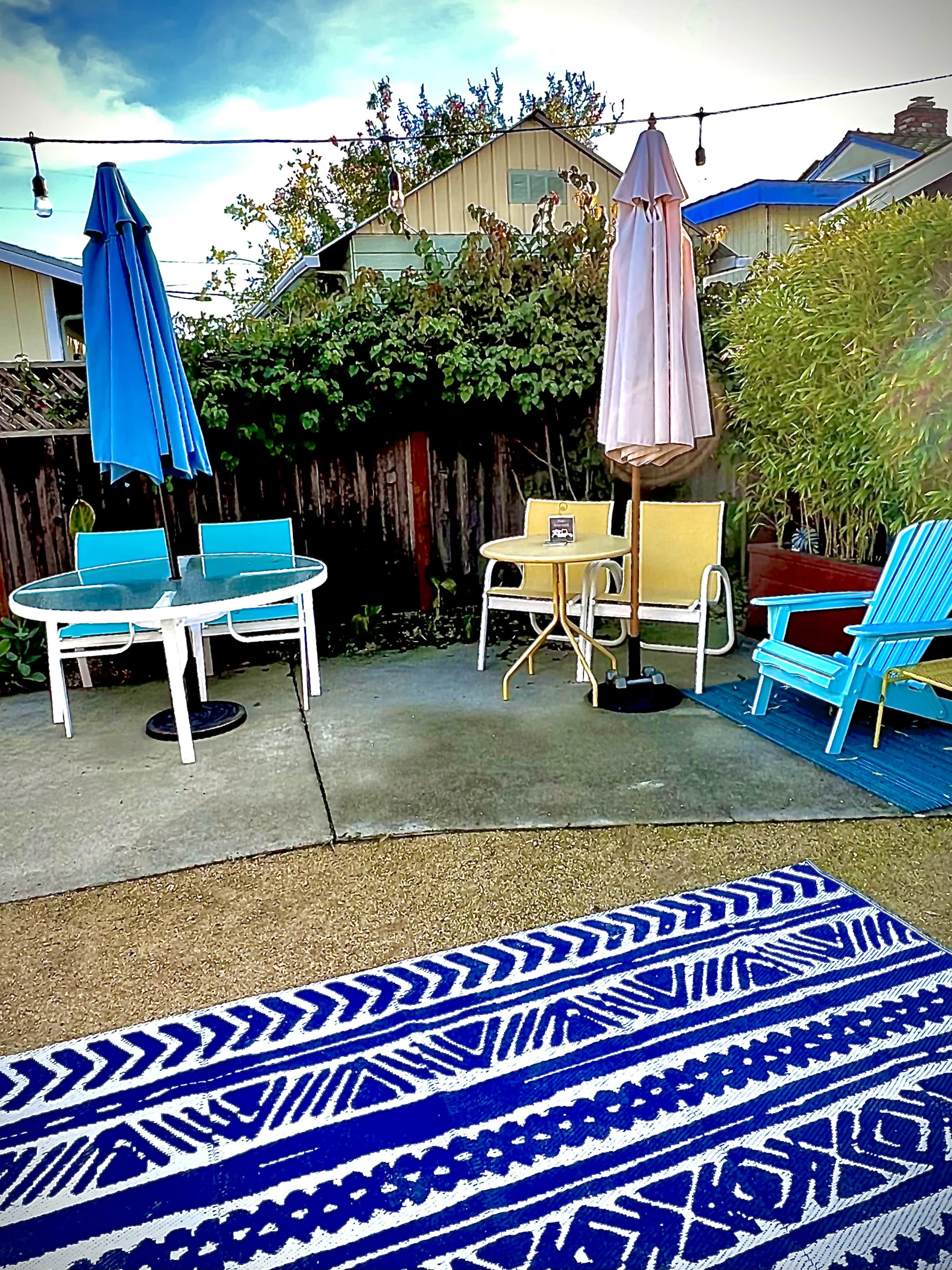 A backyard scene with various chairs and tables arranged on a concrete patio, surrounded by greenery and decorative lighting.