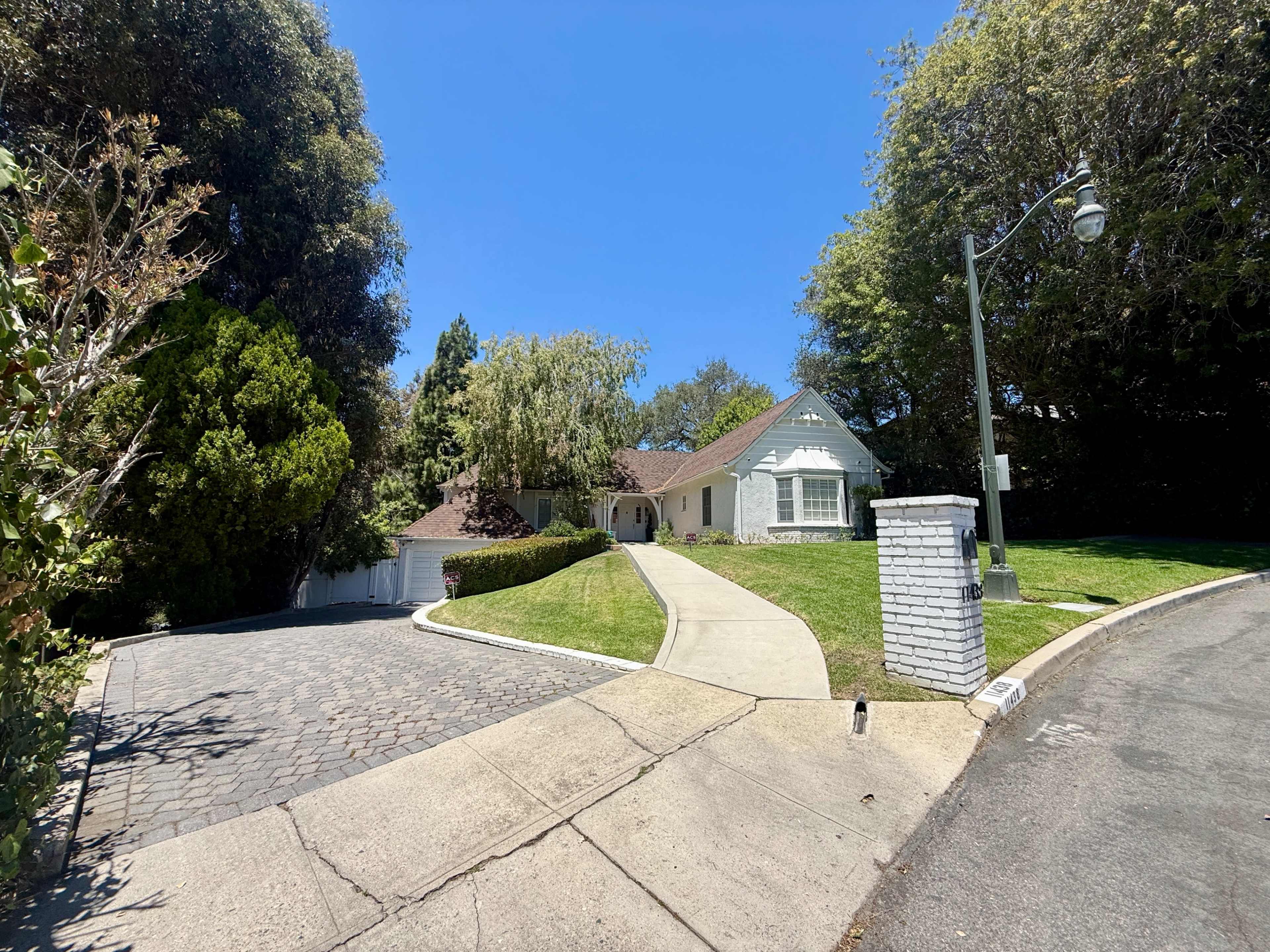 The image shows a curved driveway leading to a single-story house surrounded by lush greenery on a sunny day.