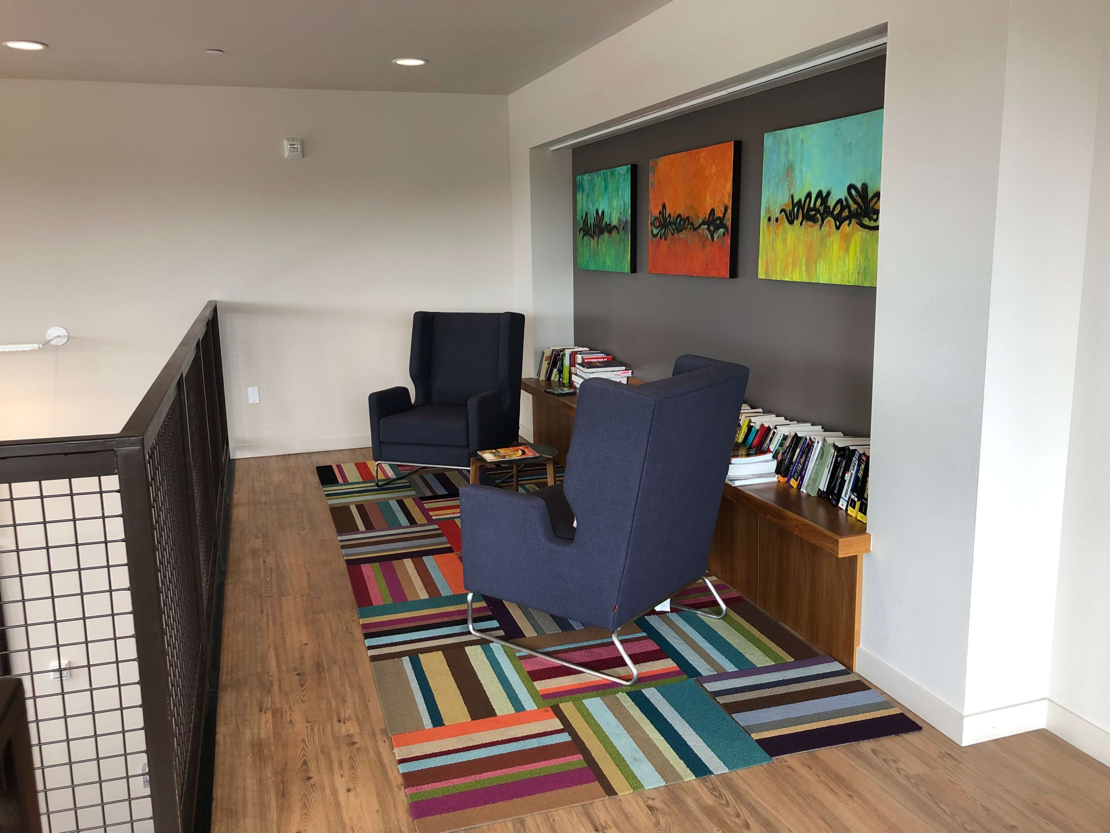 The image shows a modern seating area with two blue chairs, a colorful striped rug, and shelves filled with books against a light-colored wall.