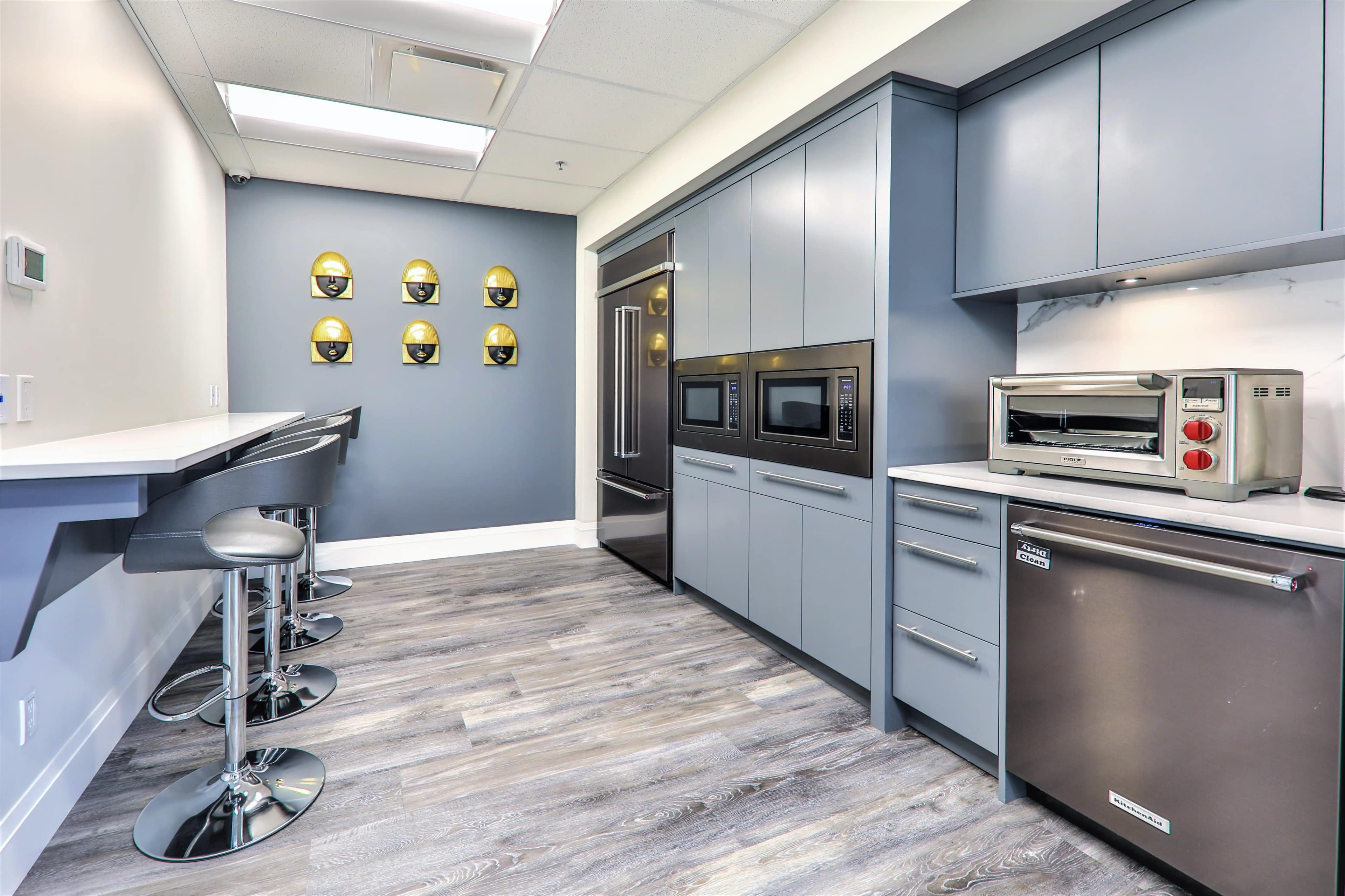 The image shows a modern kitchen with gray cabinetry, stainless steel appliances, and a breakfast bar with circular stools.