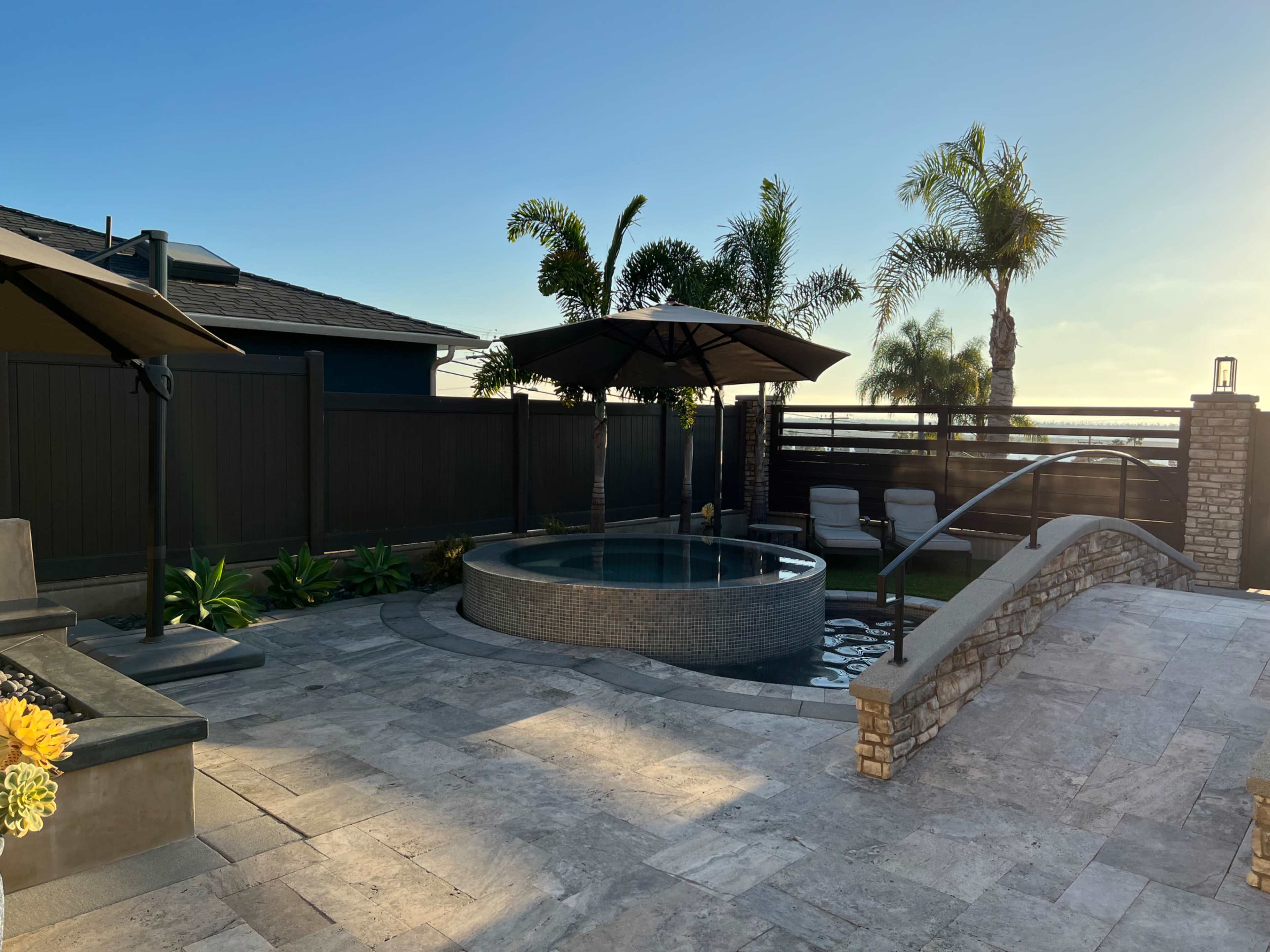 A backyard with a circular hot tub, two lounge chairs under large umbrellas, and palm trees against a clear sky.