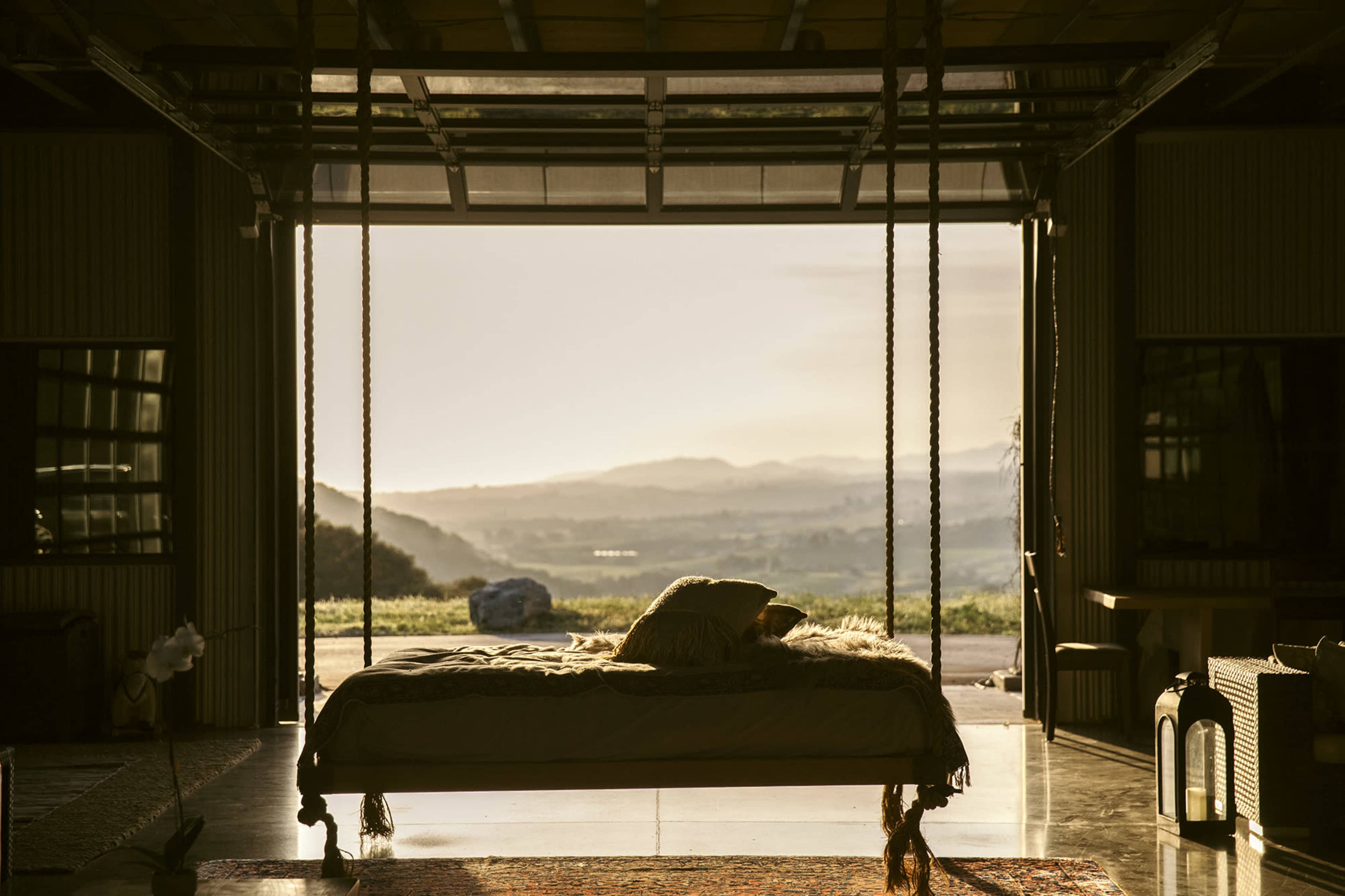 A bed with soft bedding is suspended from the ceiling in an open garage-like space that looks out onto a distant landscape at sunset.