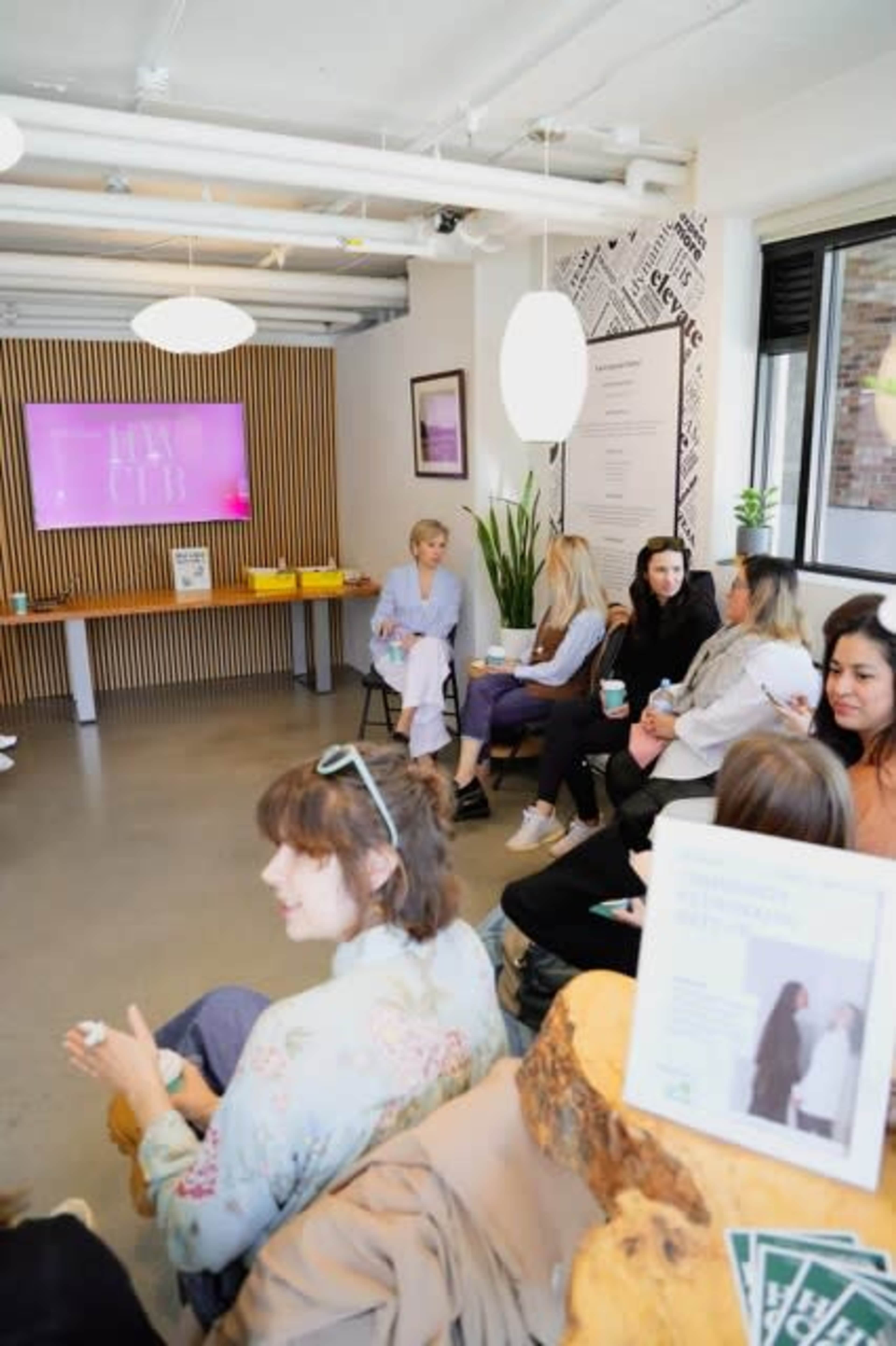 A group of people is seated in a modern, well-lit room with a presentation screen displaying "HUB," while a few individuals converse and hold drinks.
