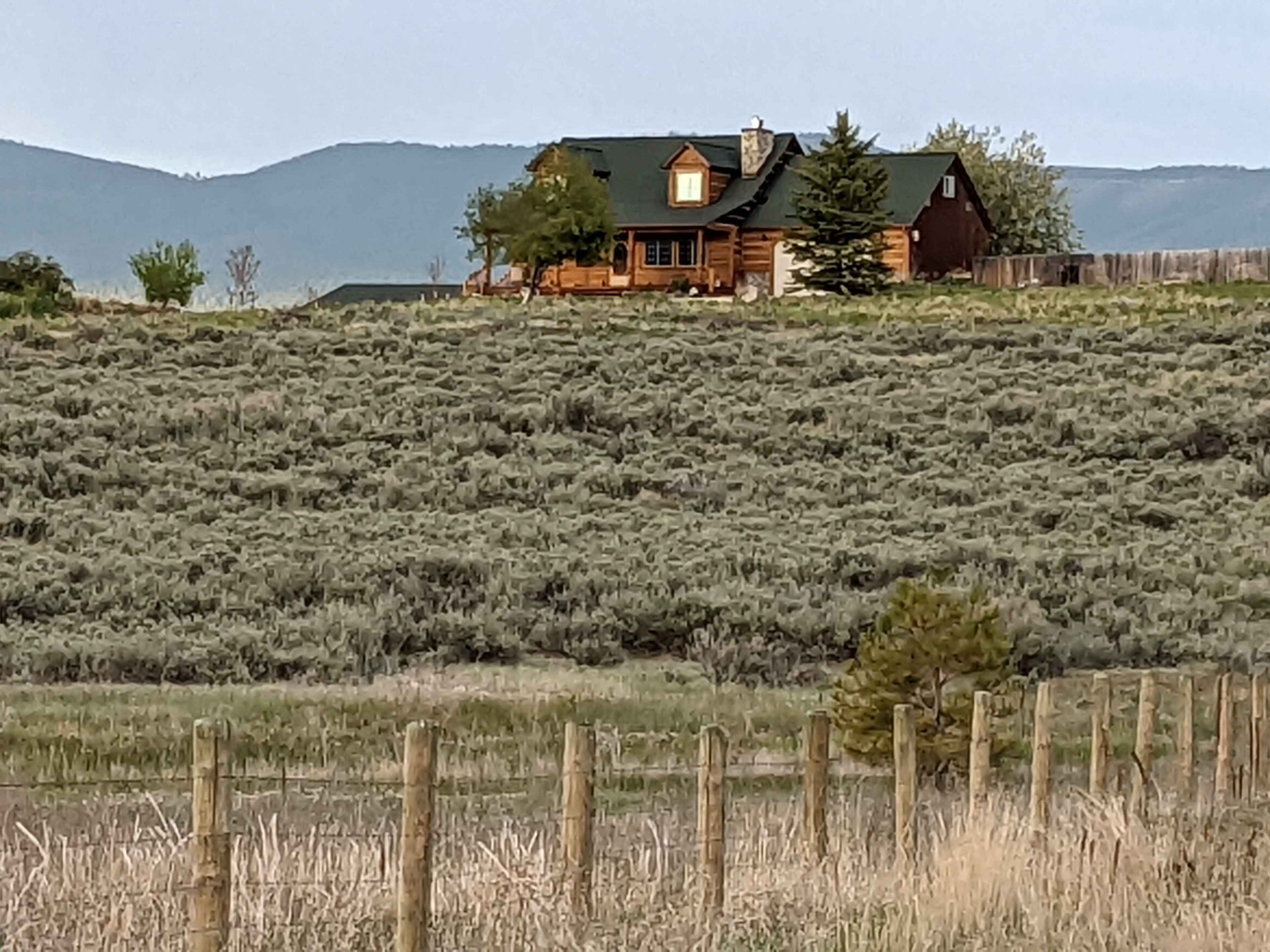 A log cabin with a green roof is situated on a hillside surrounded by sagebrush and mountains in the background.