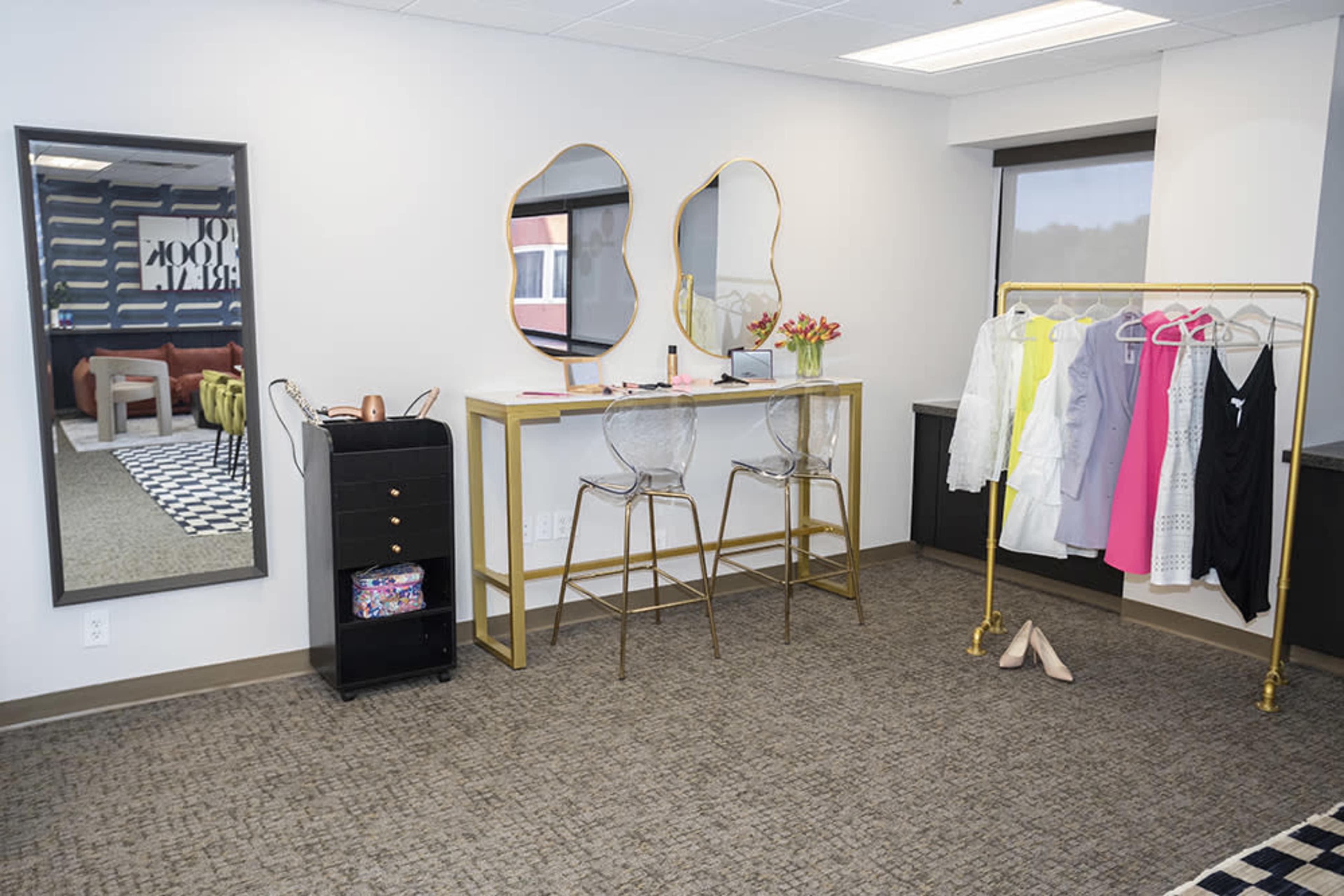 The image shows a modern interior space featuring a gold-accented table with two transparent chairs, a clothing rack with brightly colored garments, and a full-length mirror against a light-colored wall.