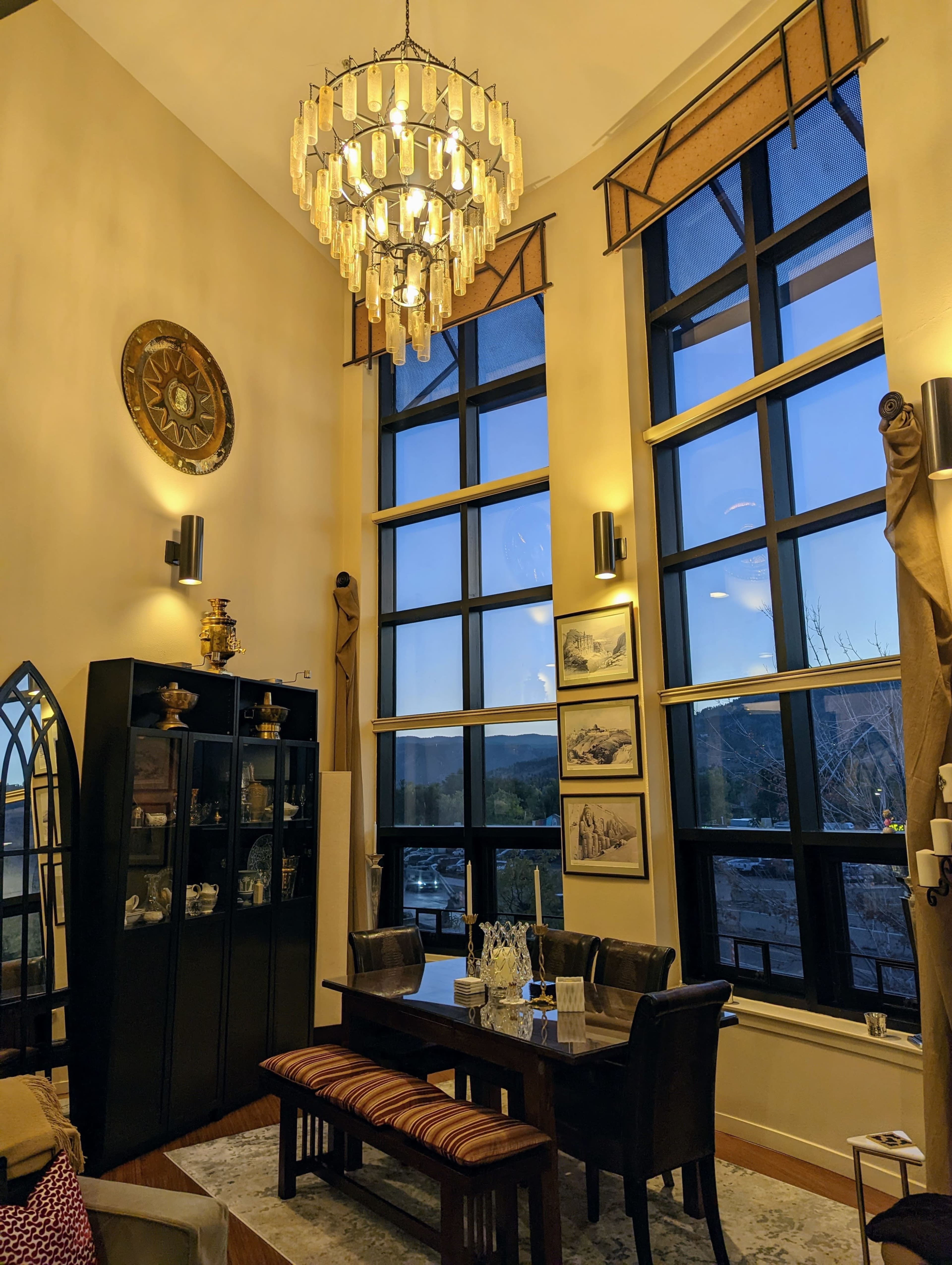 A tall room features large windows, a chandelier, a dining table with chairs, and a display cabinet against the wall.