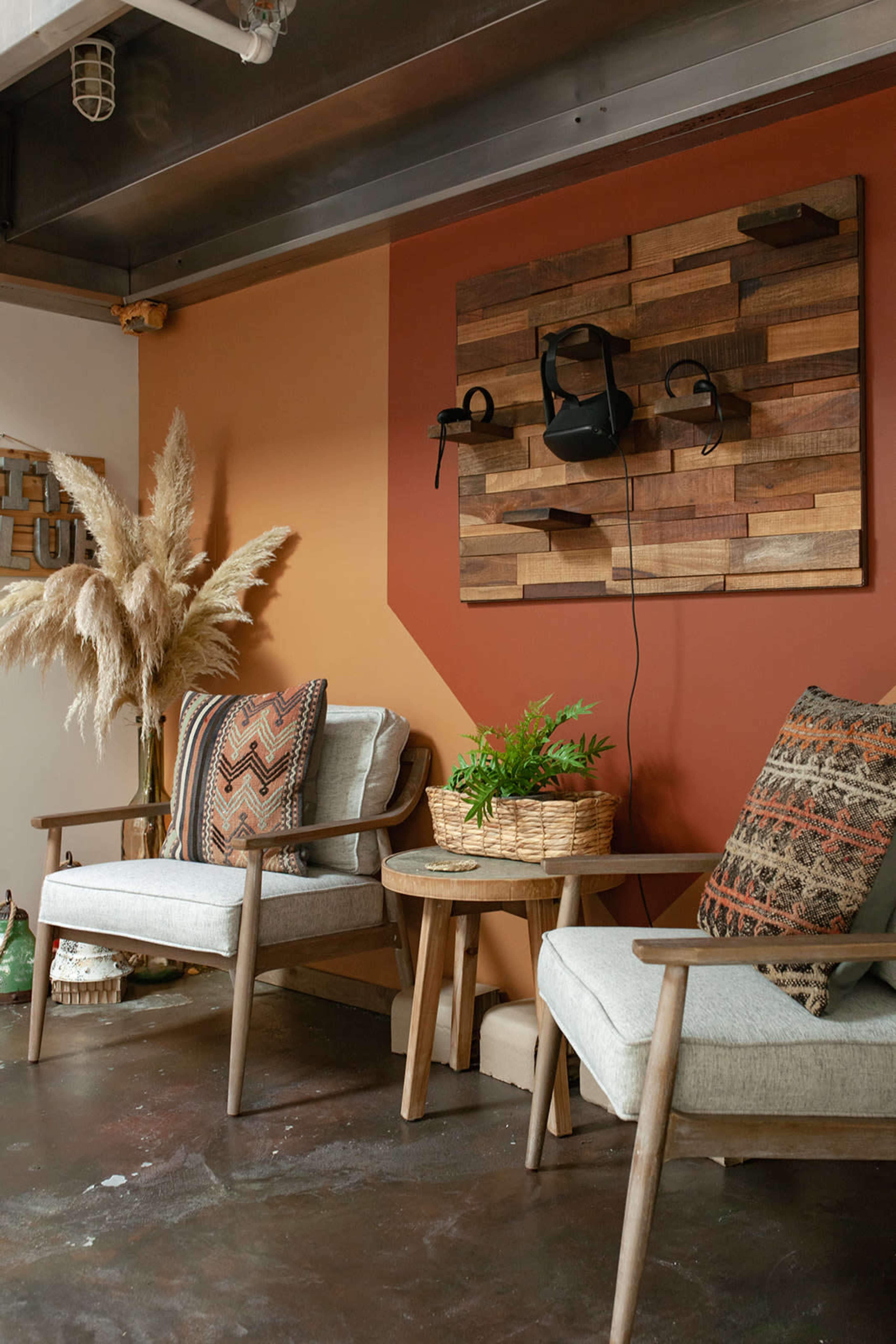 A cozy seating area features two wooden chairs with patterned cushions, a small round table, and decorative elements like a potted plant and wall art made of wooden pieces.