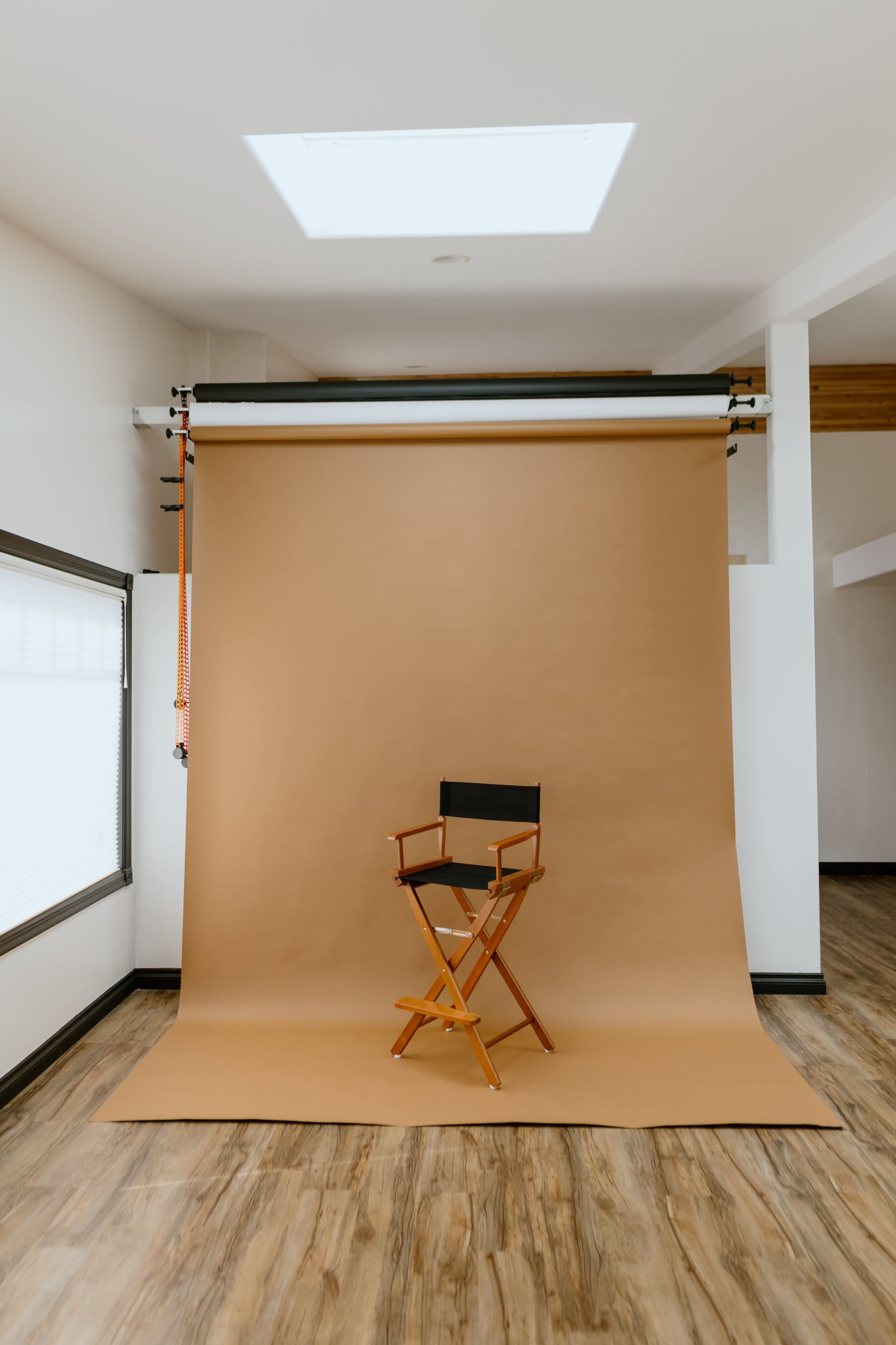 A black director's chair is positioned in front of a large, brown backdrop in a well-lit studio space with wooden flooring.