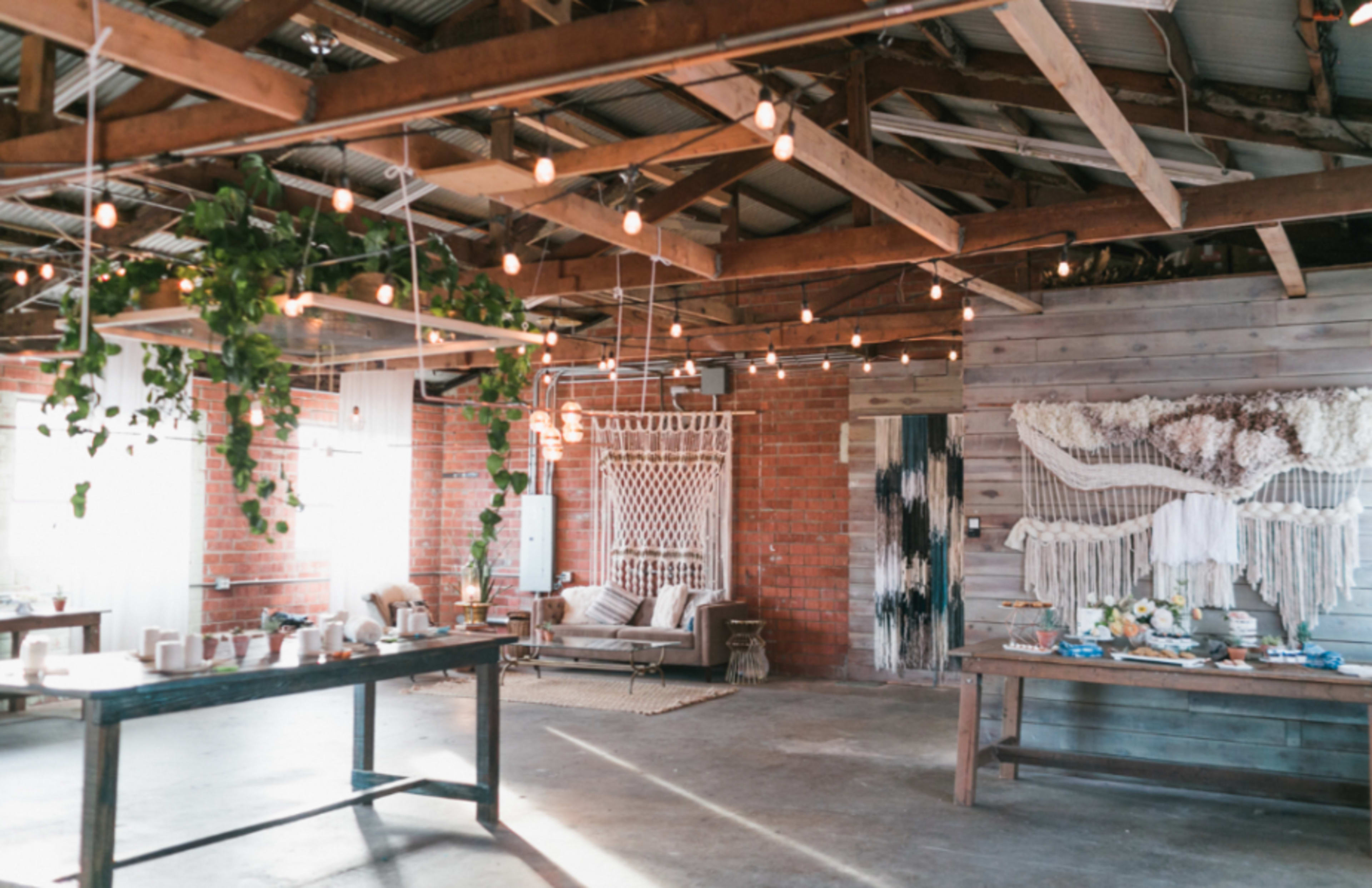 The image shows a spacious, rustic interior with wooden beams, string lights, and various decorative elements, including plants and textile art.