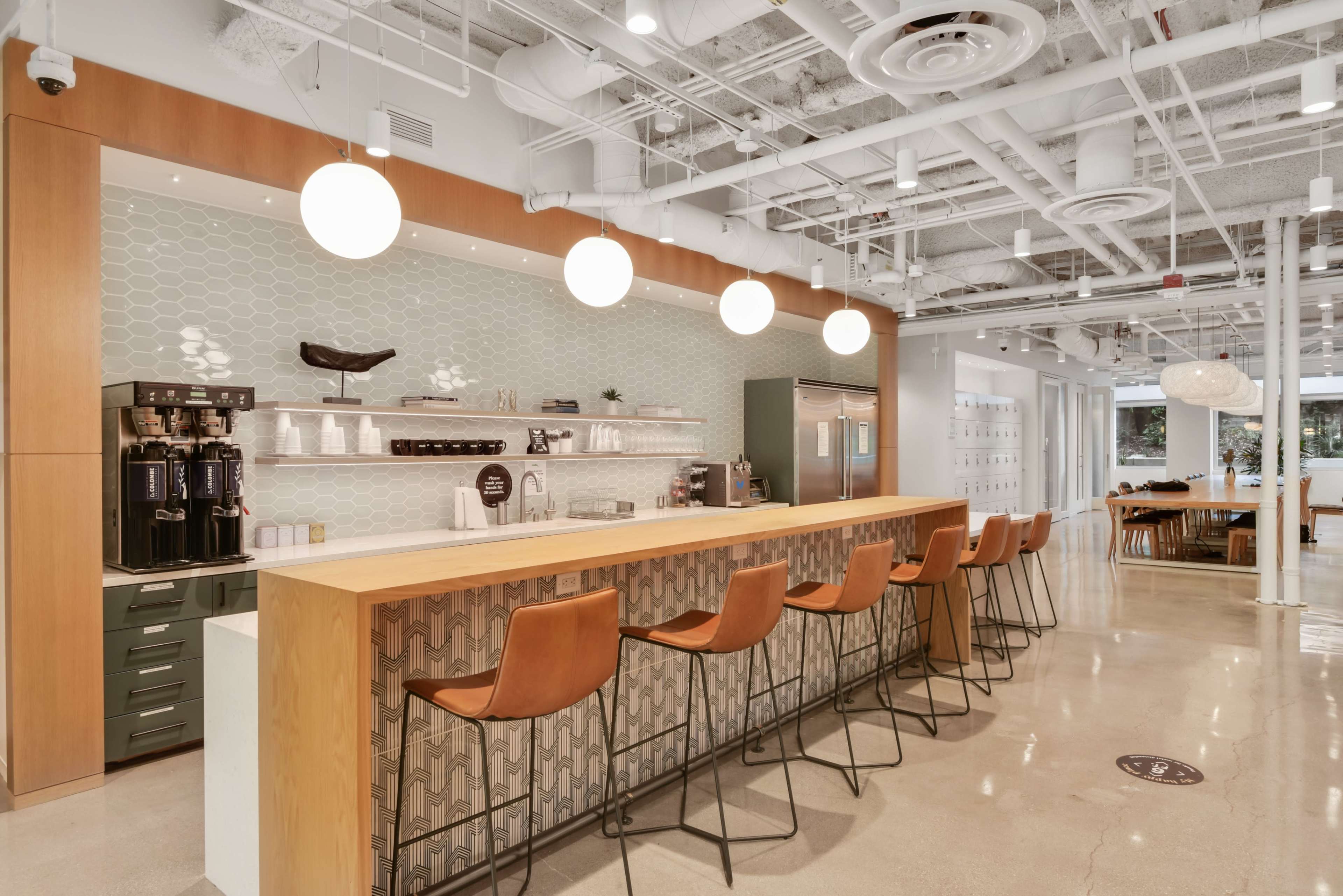 The image shows a modern office kitchen area with a long wooden bar, brown stools, and coffee machines against a patterned backsplash.